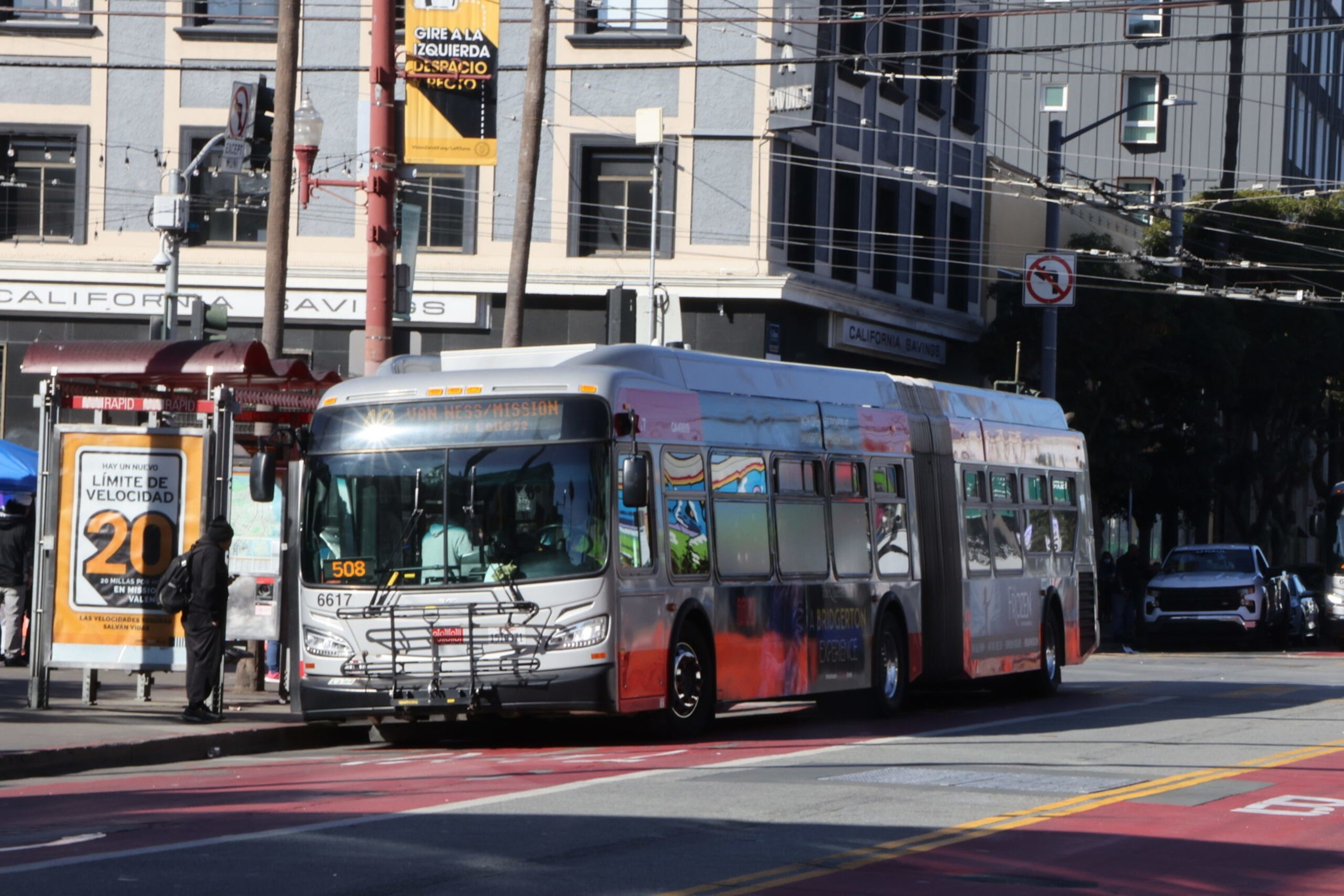A silver articulated city bus stopped at a bus stop on a sunny urban street with a 20 mph speed limit sign in Spanish nearby.