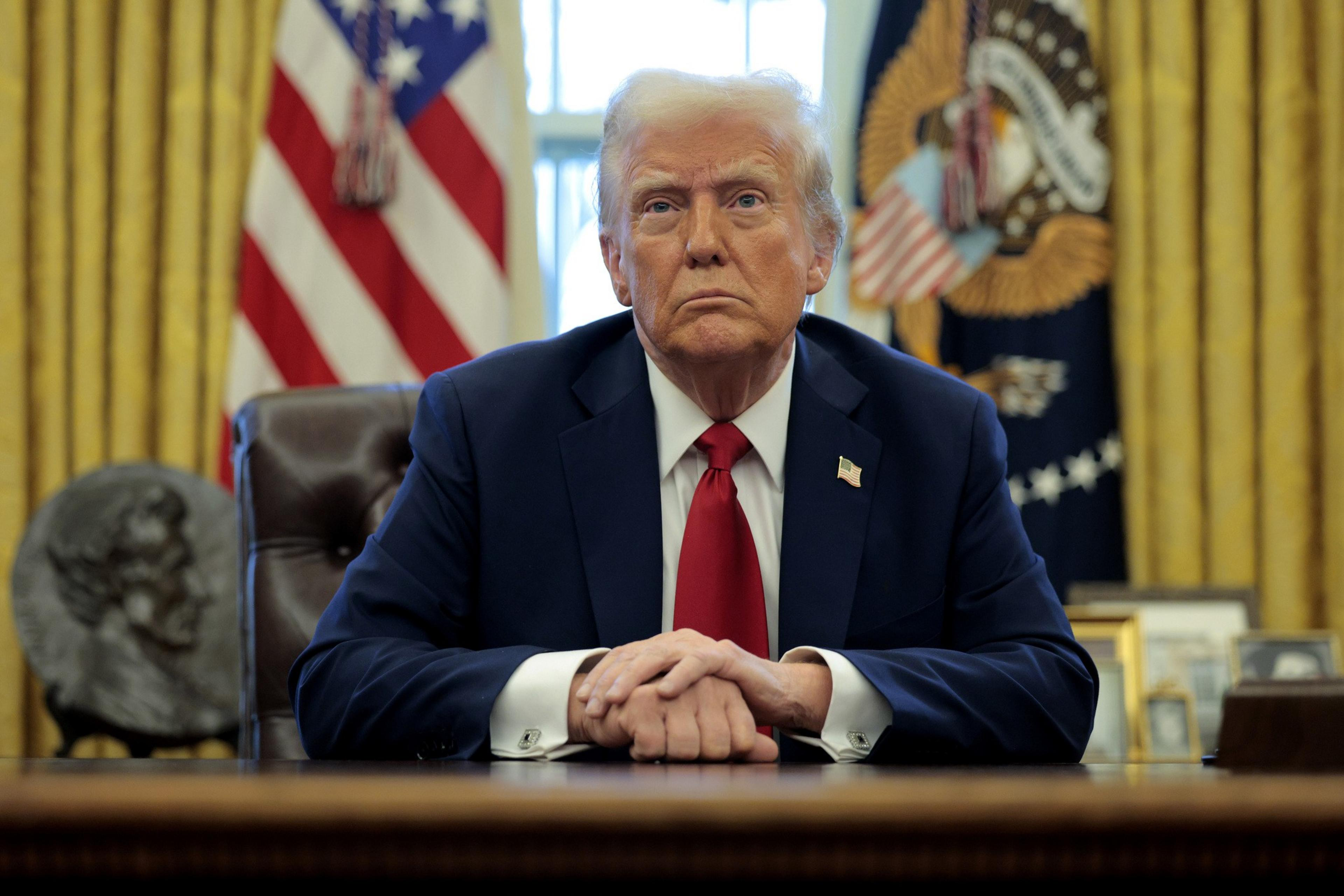 A man in a suit and red tie sits at a desk with hands clasped. Behind are American flags and a seal on gold curtains. A bust and framed photos are visible.