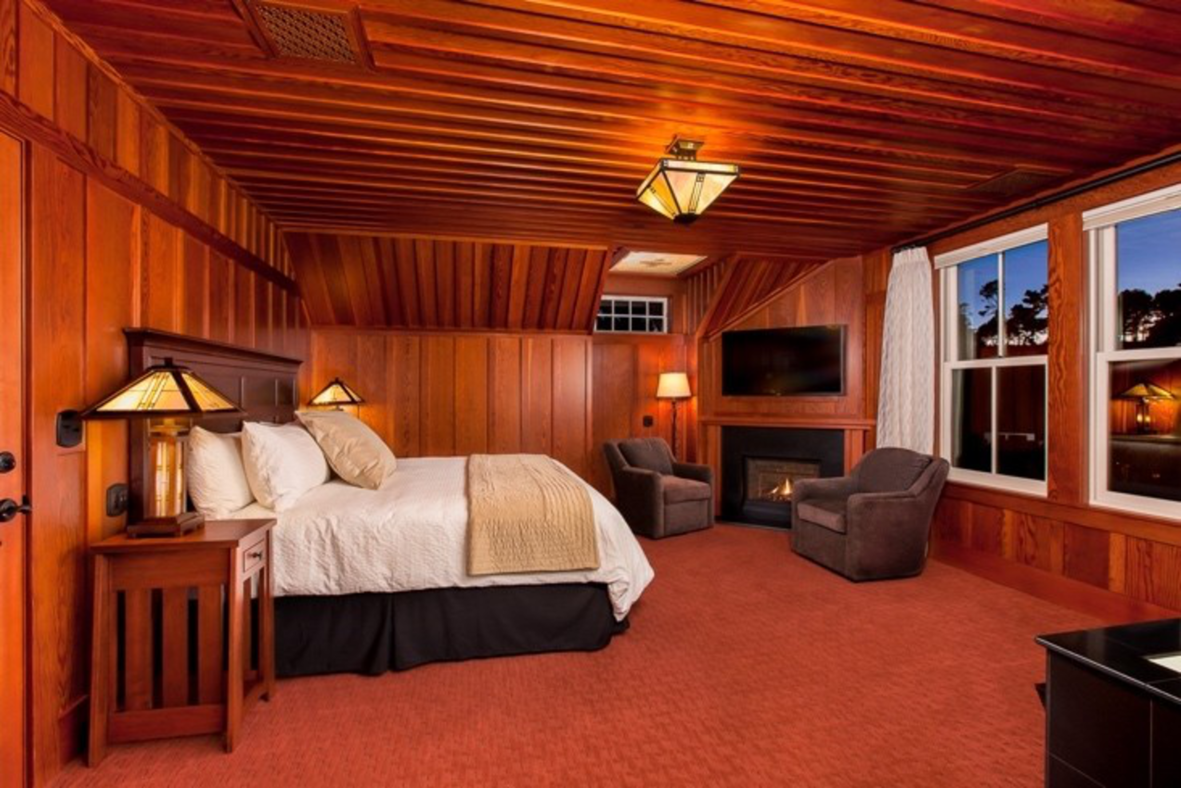 A cozy bedroom with wooden walls and ceiling, a large bed, two armchairs by a lit fireplace, a wall-mounted TV, and large windows showing a darkening sky.