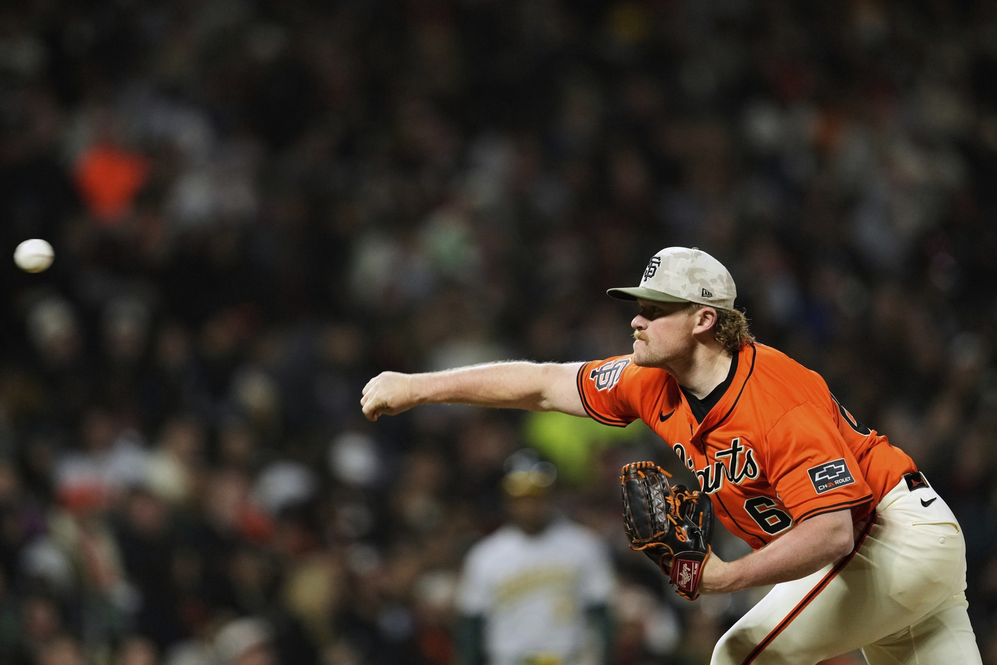 A baseball player in an orange jersey and cap is pitching the ball. The crowd in the background is blurred, and a batsman stands in focus.