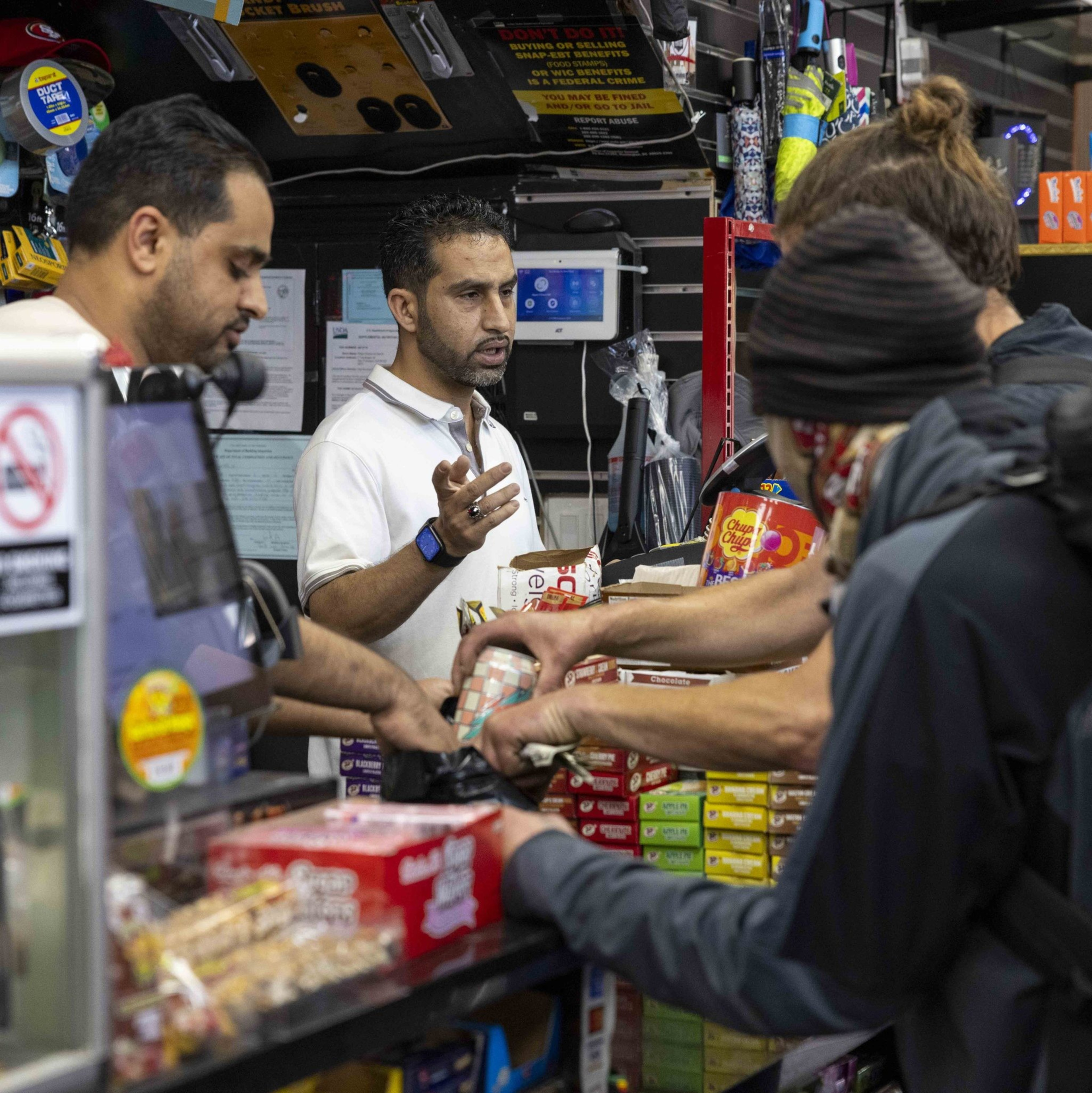 A busy store counter with two employees assisting customers. Various snacks and small items are displayed, and people are exchanging money and goods.