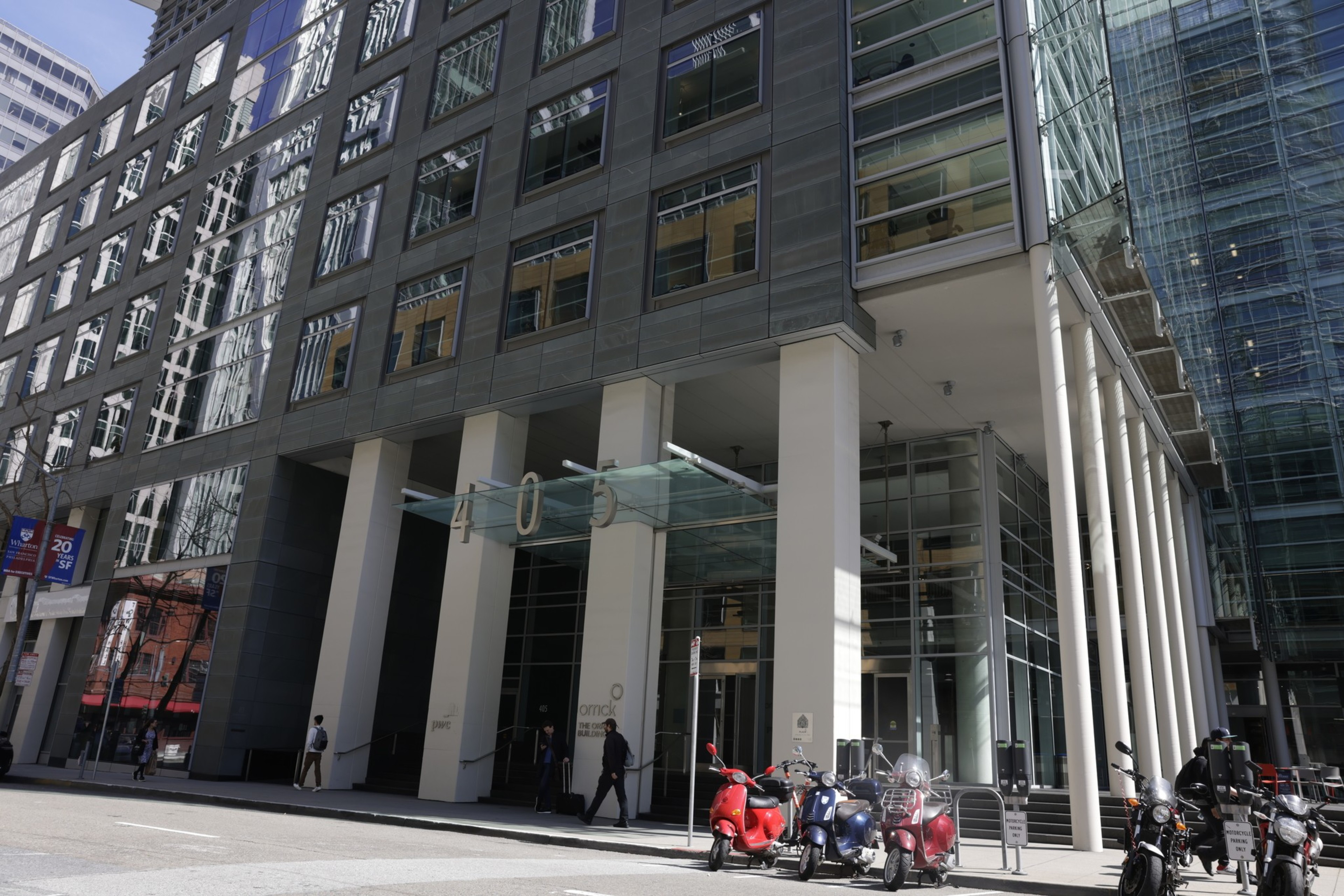 A modern building facade with large glass doors, tall columns, and parked scooters in front.