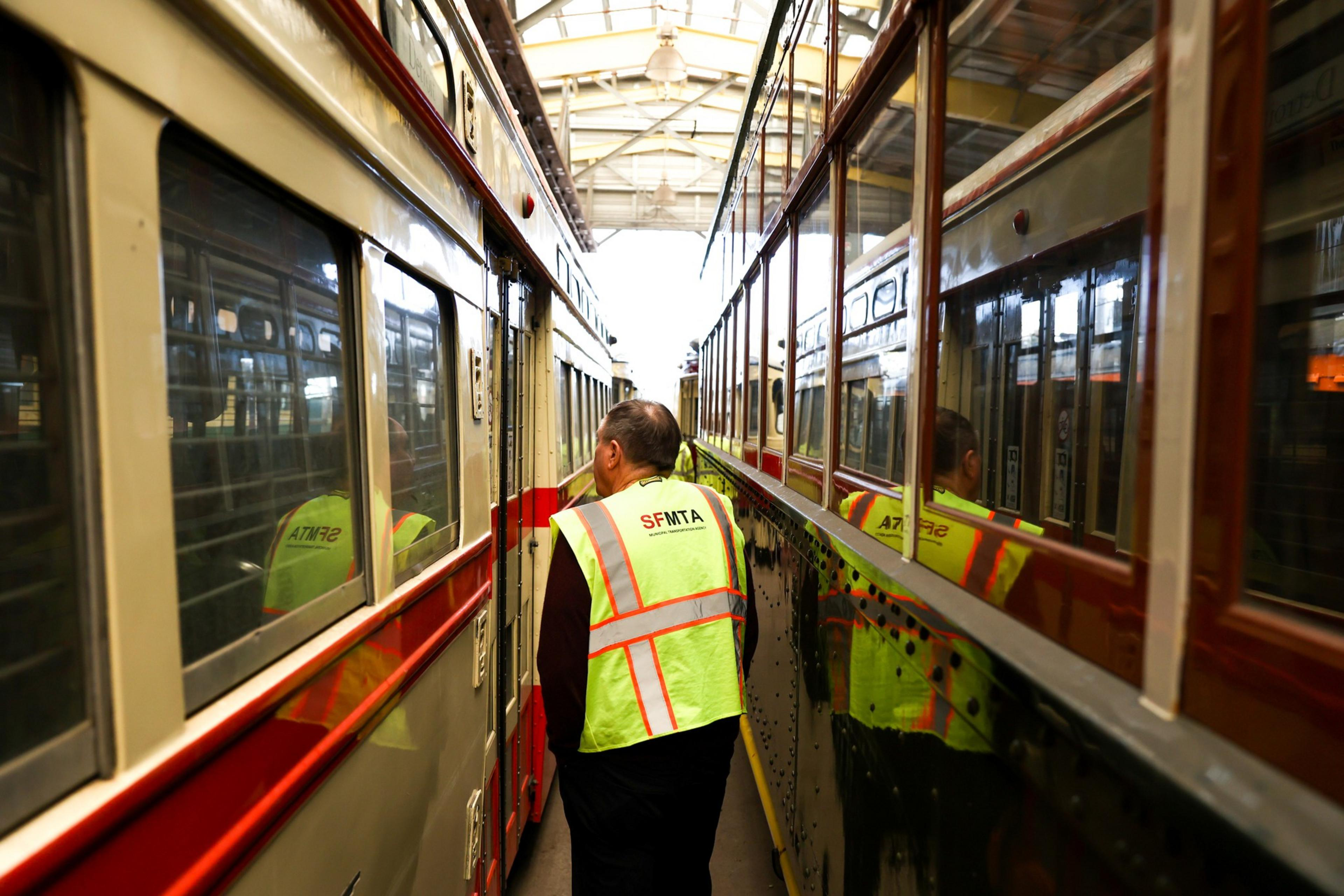 A person wearing a yellow safety vest labeled "SFMTA" walks between two close, red-and-cream streetcars inside a covered depot.