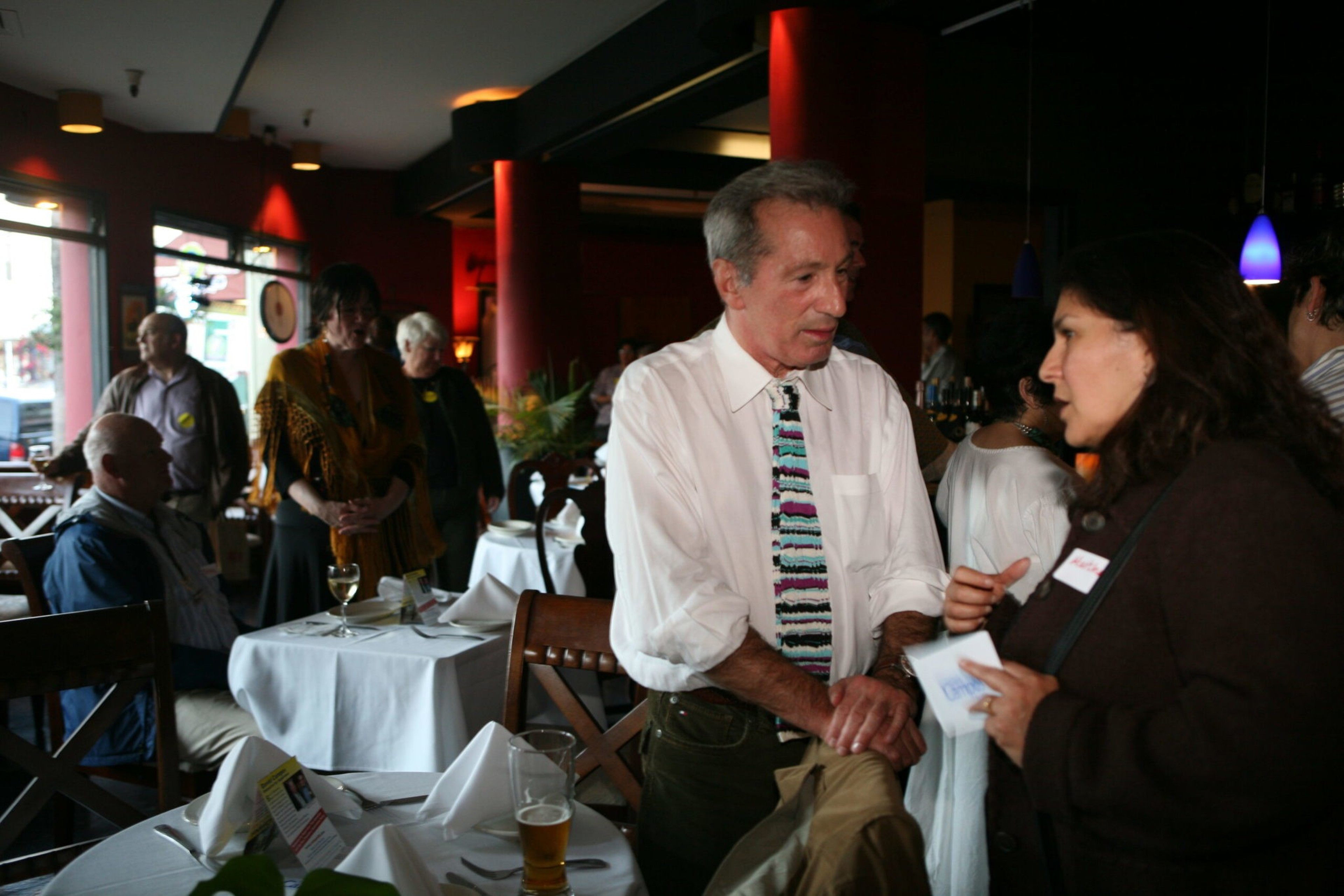 A man in a white shirt and colorful tie talks to a woman wearing a dark coat in a dimly lit restaurant with other seated and standing guests.