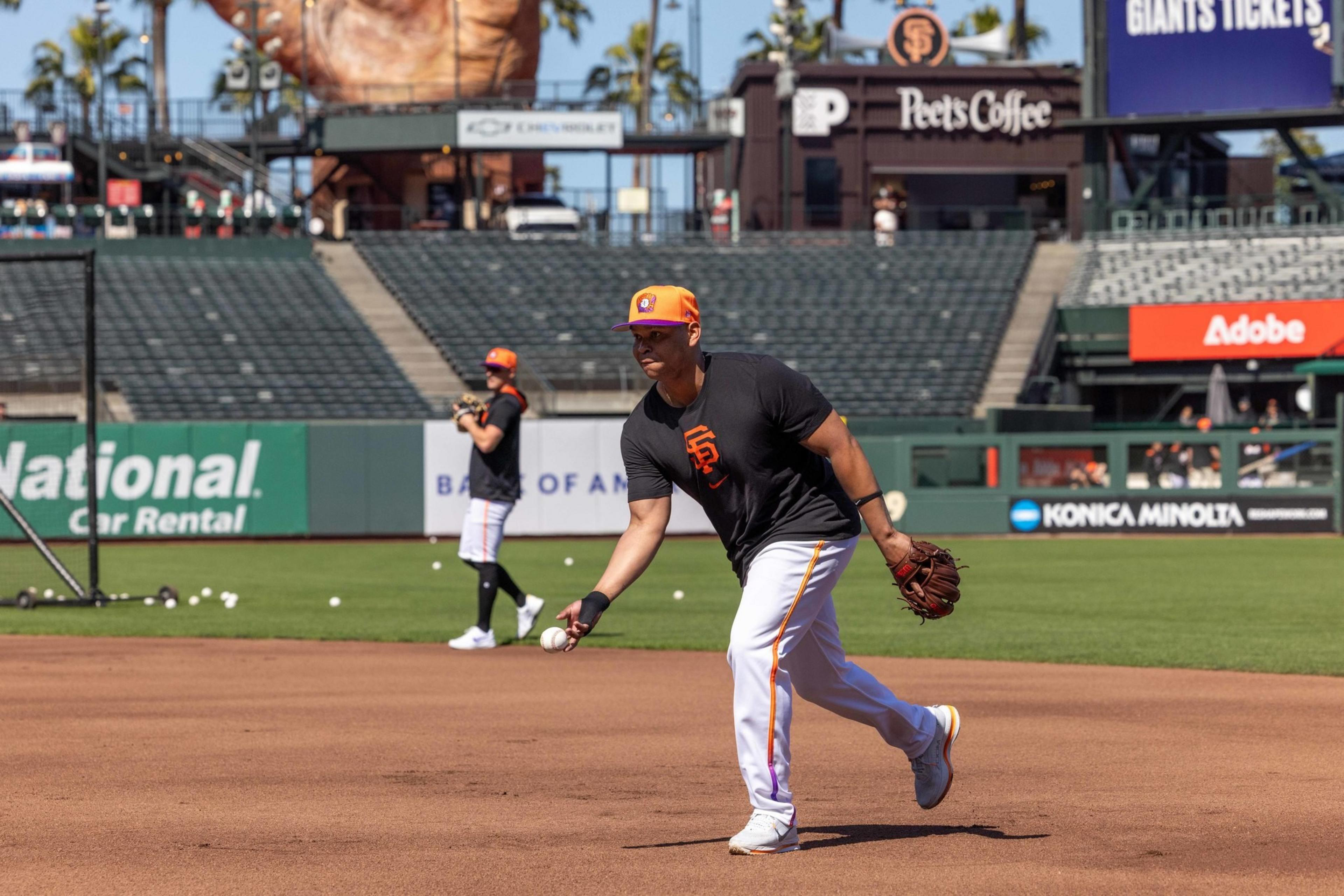 Two baseball players in orange caps and black shirts practice on a field, with one throwing a ball. The background shows empty stands and various advertisements.