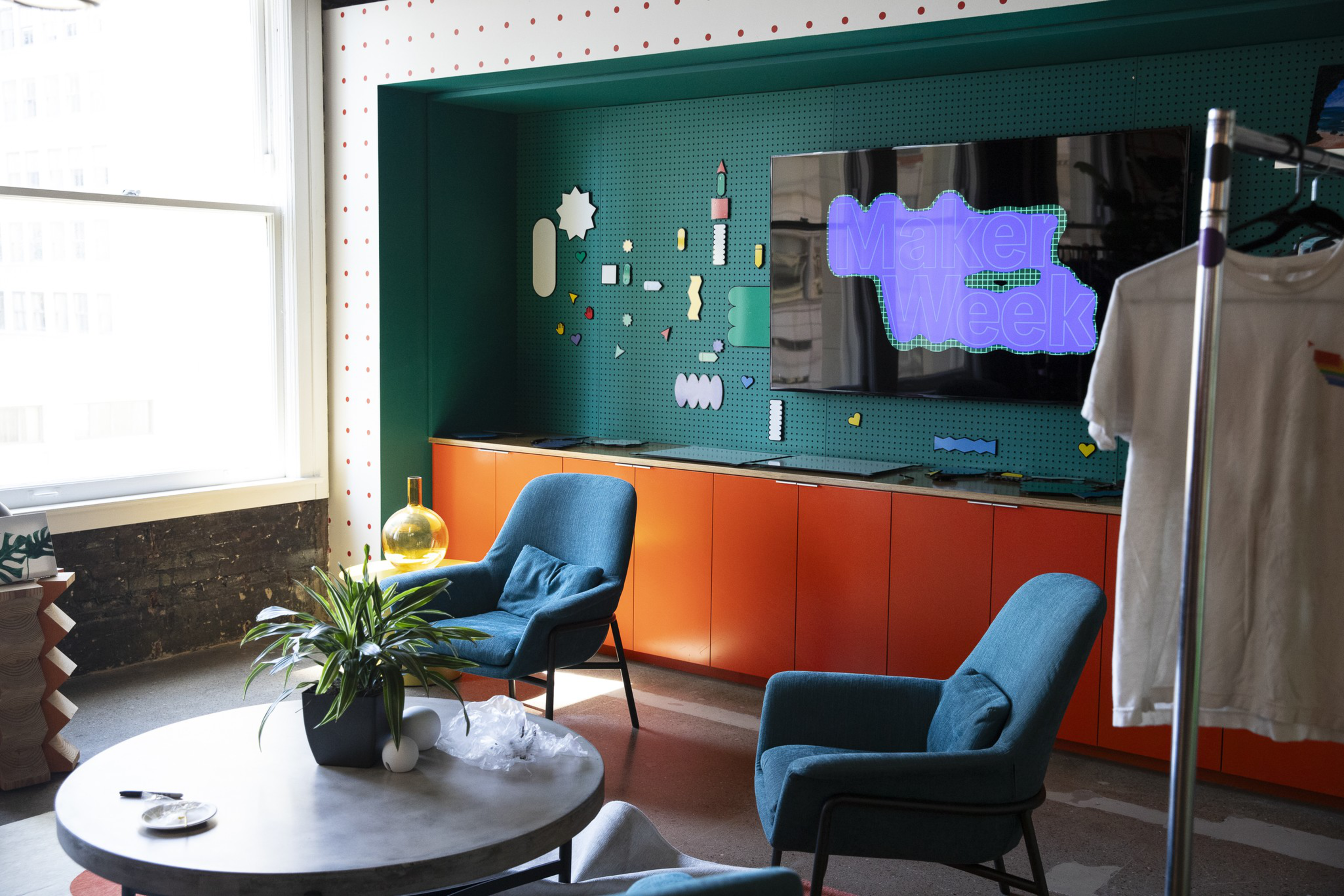 Two blue armchairs face a round table with a plant, a TV on a green wall displays "Maker Week," and orange cabinets line the back wall beneath it.
