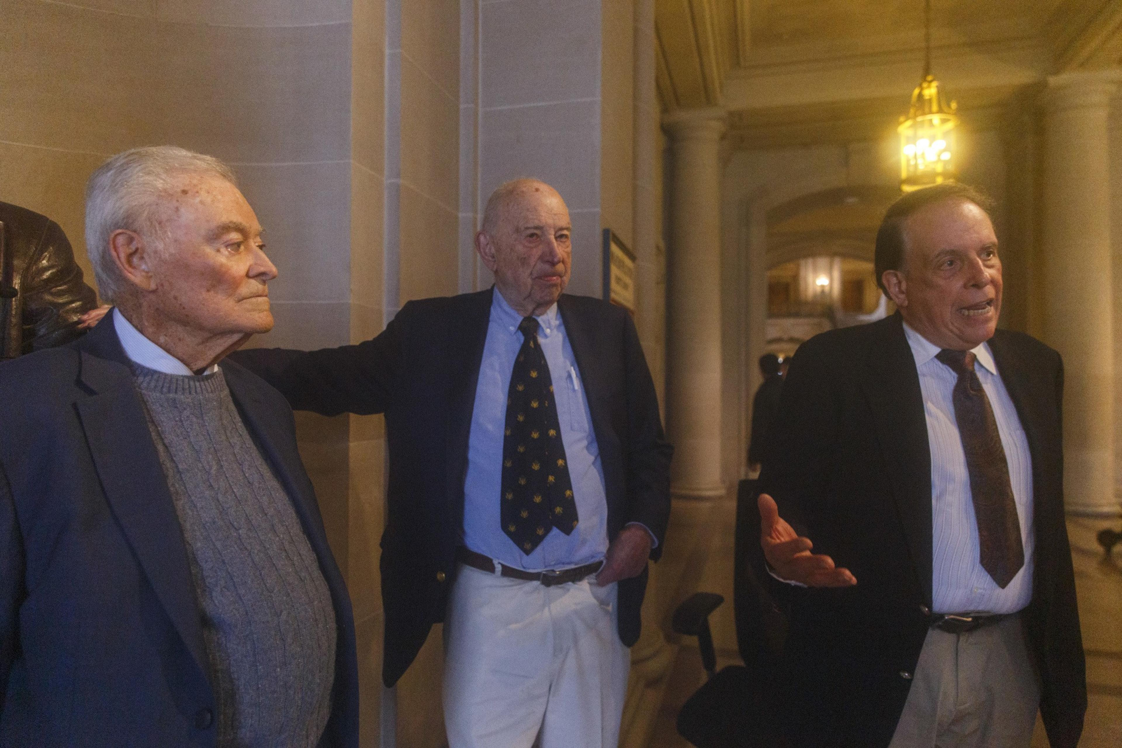 Three elderly men in suits are gathered in a room with classical architecture and warm lighting.