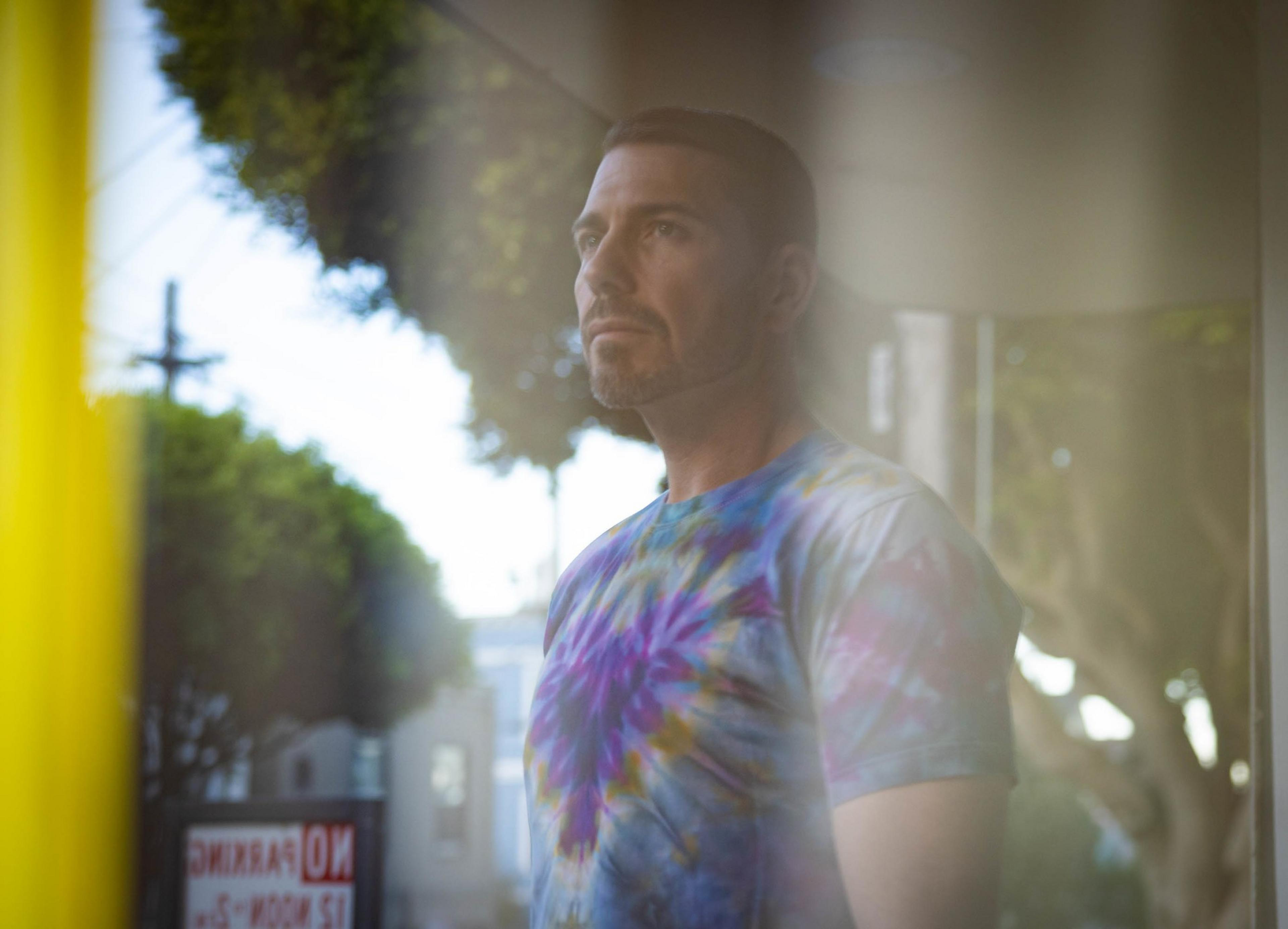A man with short hair and a beard wears a colorful tie-dye shirt, standing indoors near a window with trees and a "No Parking" sign visible outside.
