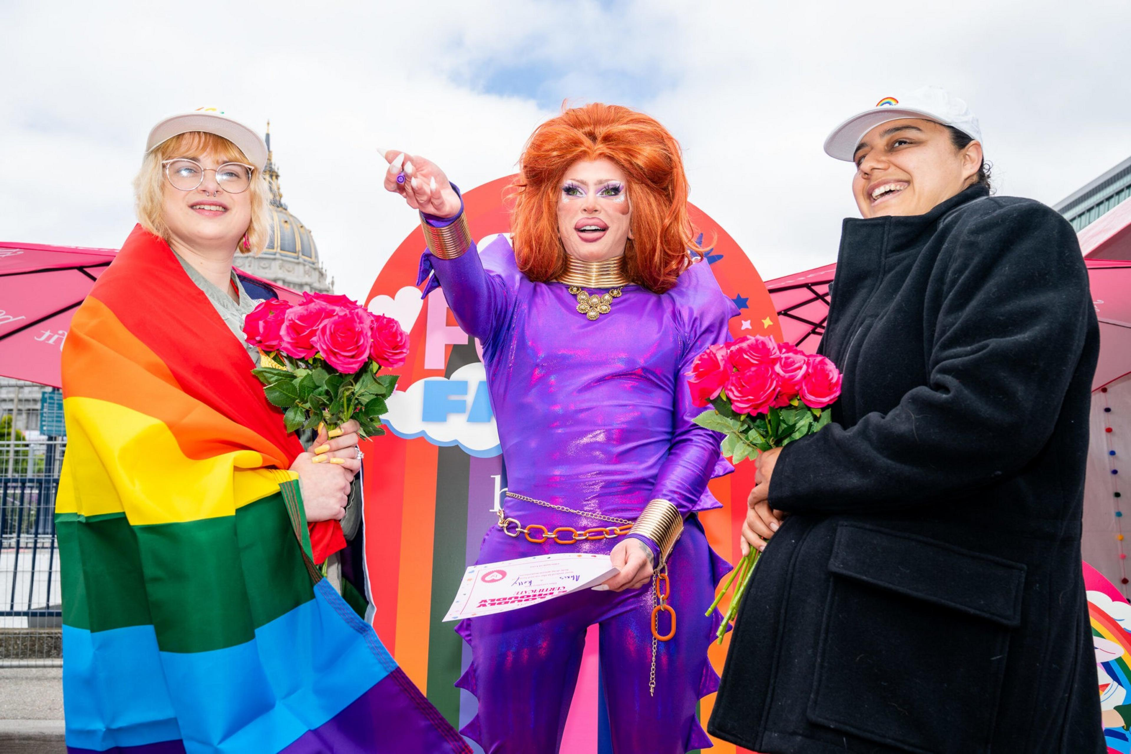 Three people smile and hold pink roses, with one in a colorful rainbow cape and another in a vibrant purple outfit and red wig.