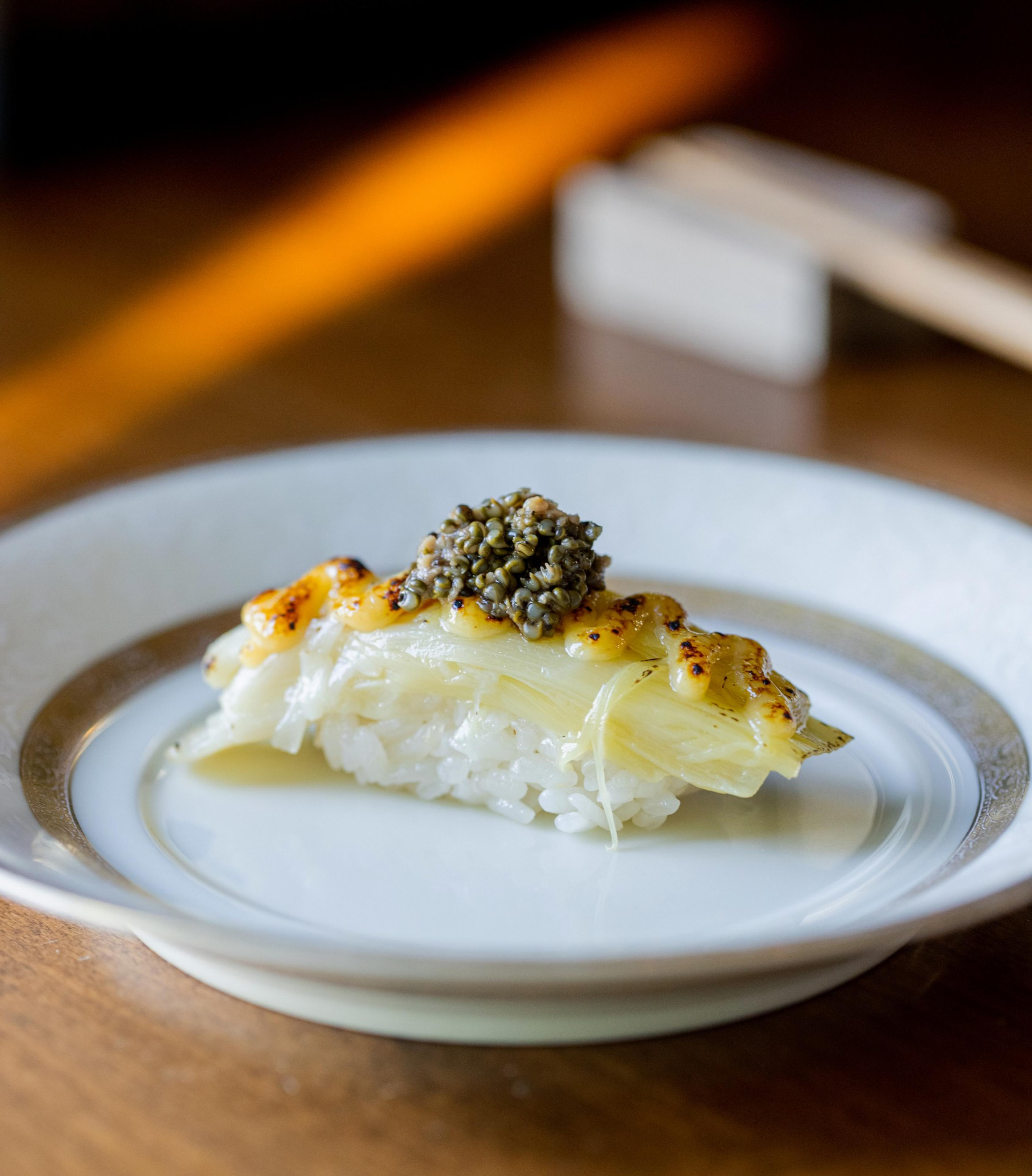 A piece of sushi topped with layers of grilled fish and a generous serving of caviar sits on a decorative white plate with gold accents.
