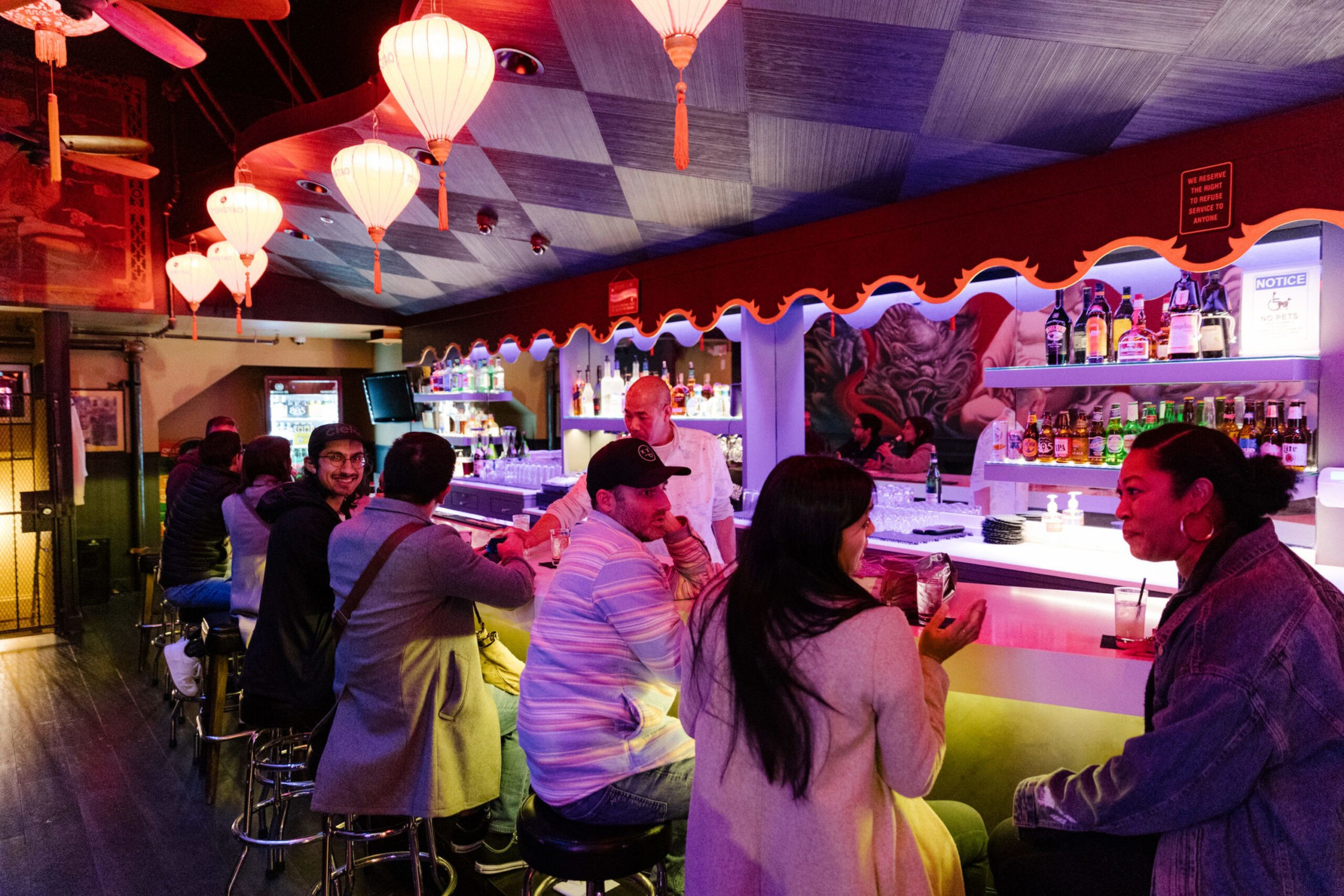 People sit and chat at a colorful bar with hanging lanterns and shelves stocked with various bottles, while a bartender stands behind the counter.