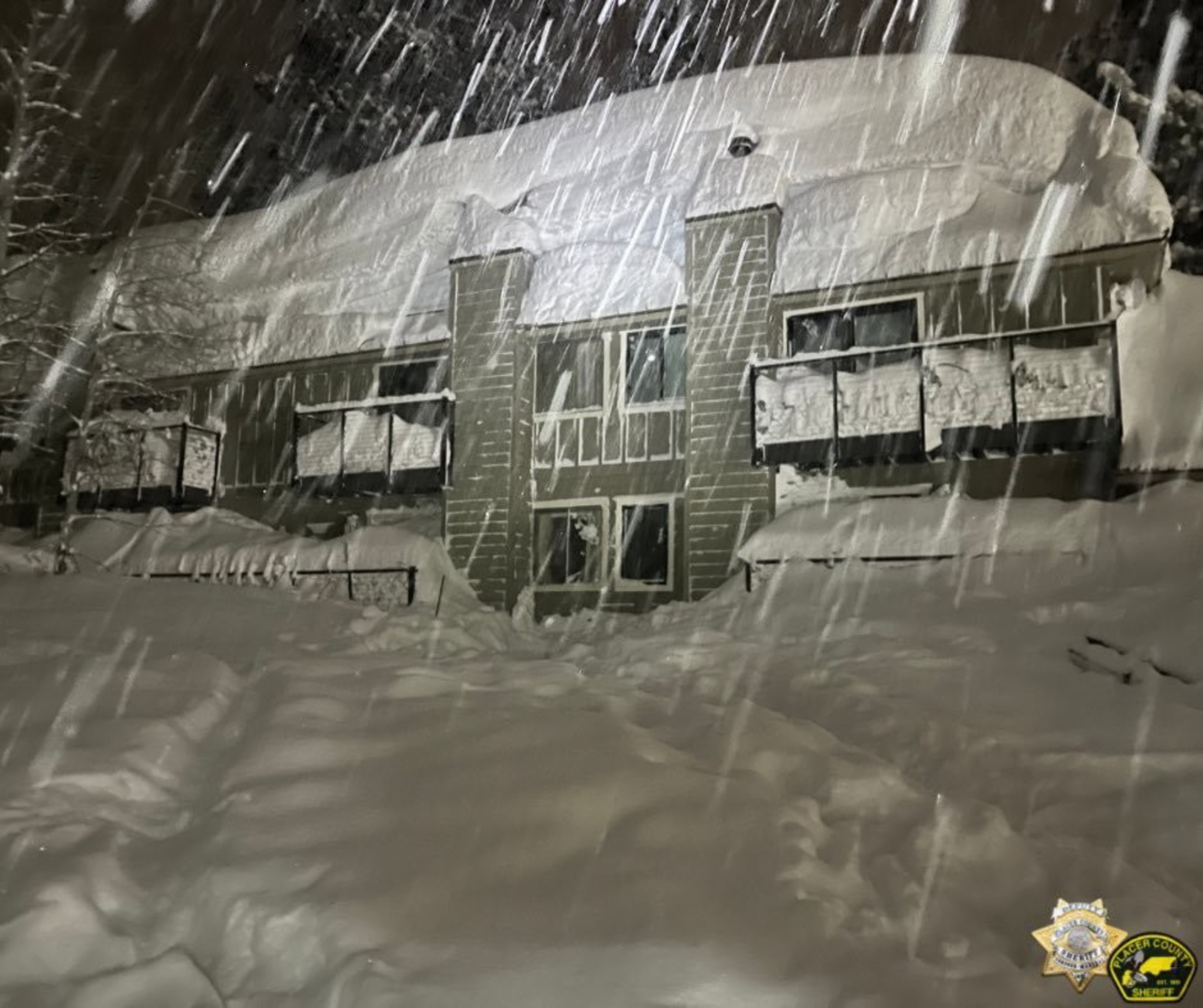 A two-story building is covered in heavy snow during a nighttime snowstorm, with thick snow piled on the roof, balconies, and ground.
