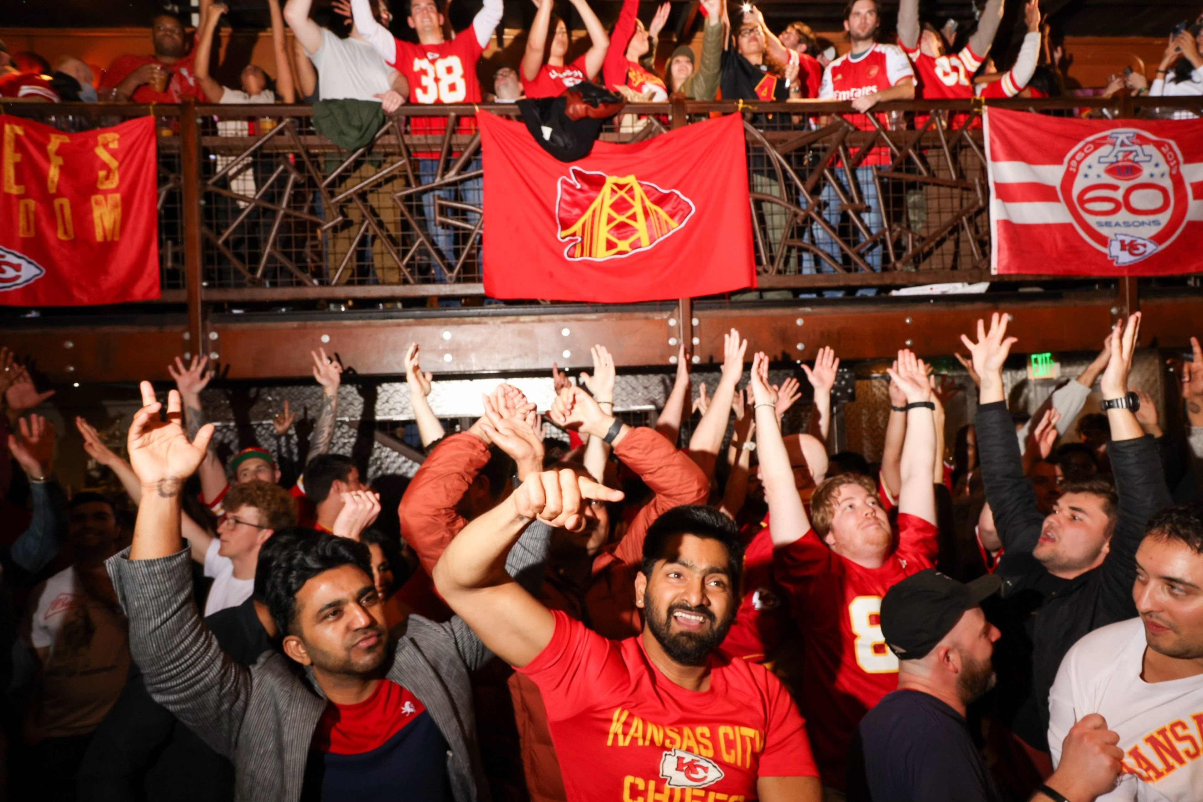 Excited fans in red apparel cheer at a sports event, with hands raised and team flags displayed.