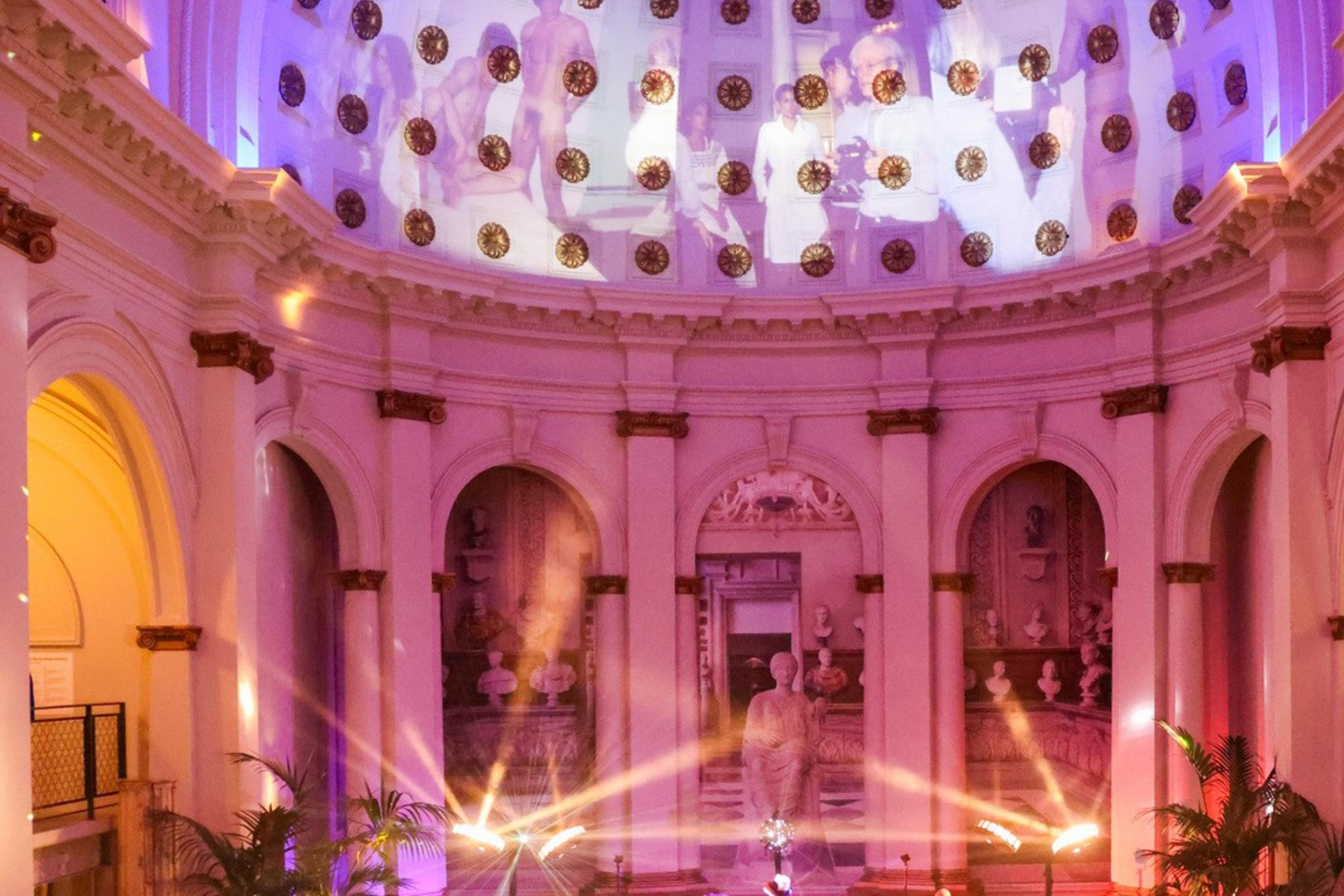 A vibrant party in a grand hall features a domed ceiling, colorful lights, and people mingling and dancing. A DJ is set up on a stage surrounded by ornate arches.
