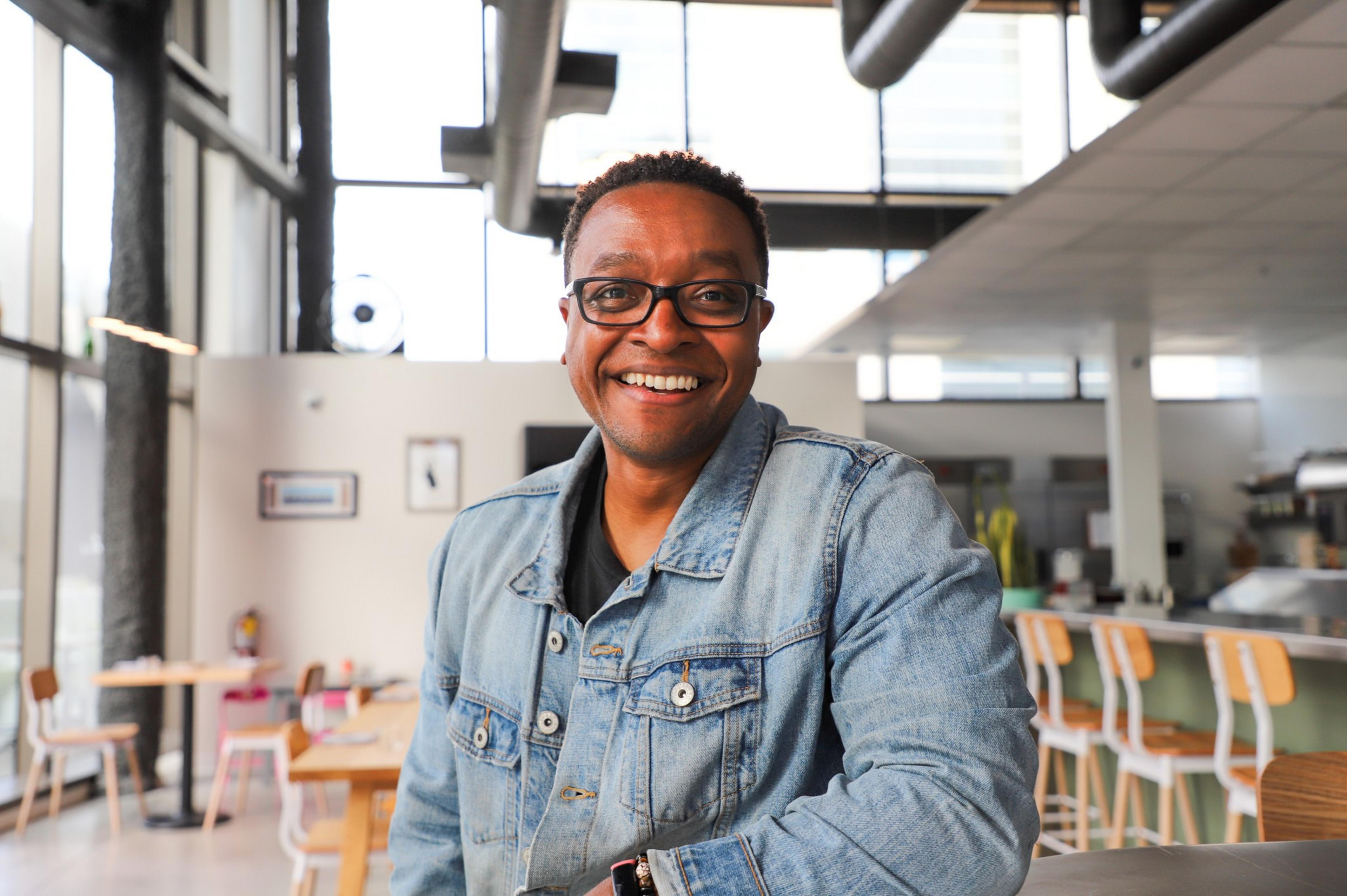 A smiling man wearing glasses and a blue denim jacket sits in a bright, modern cafe with wooden chairs and large windows.