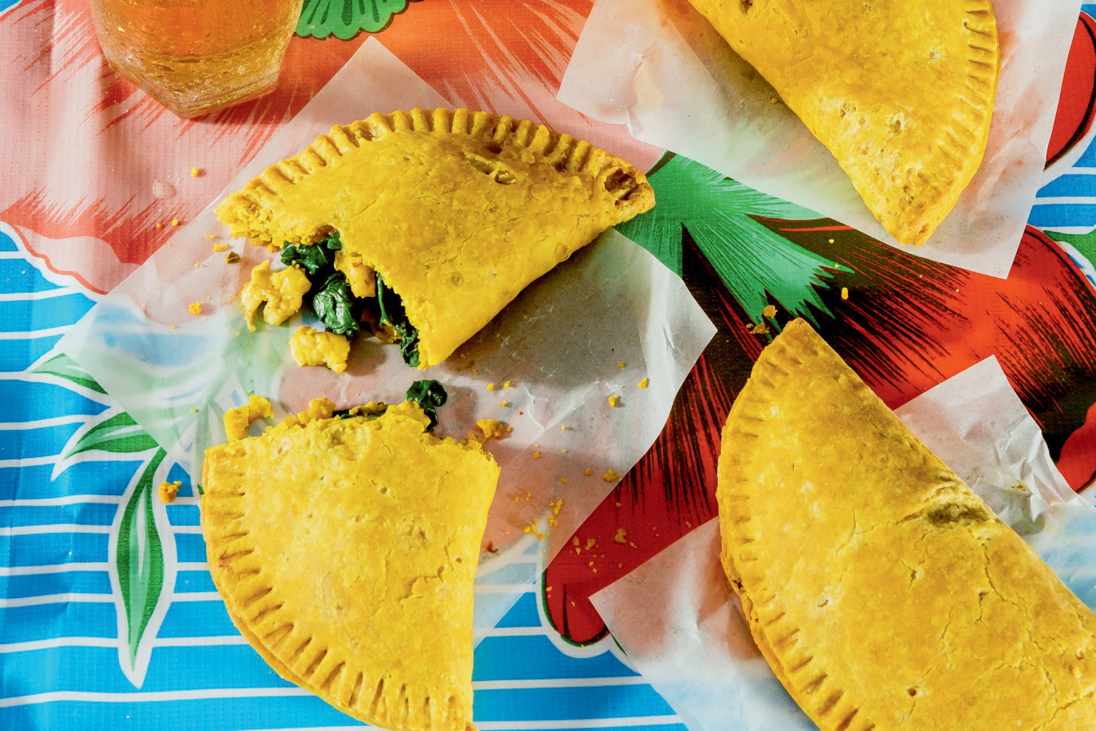 Three golden, half-moon shaped fried pastries sit on wax paper, one broken open to reveal a green leafy vegetable and egg filling.