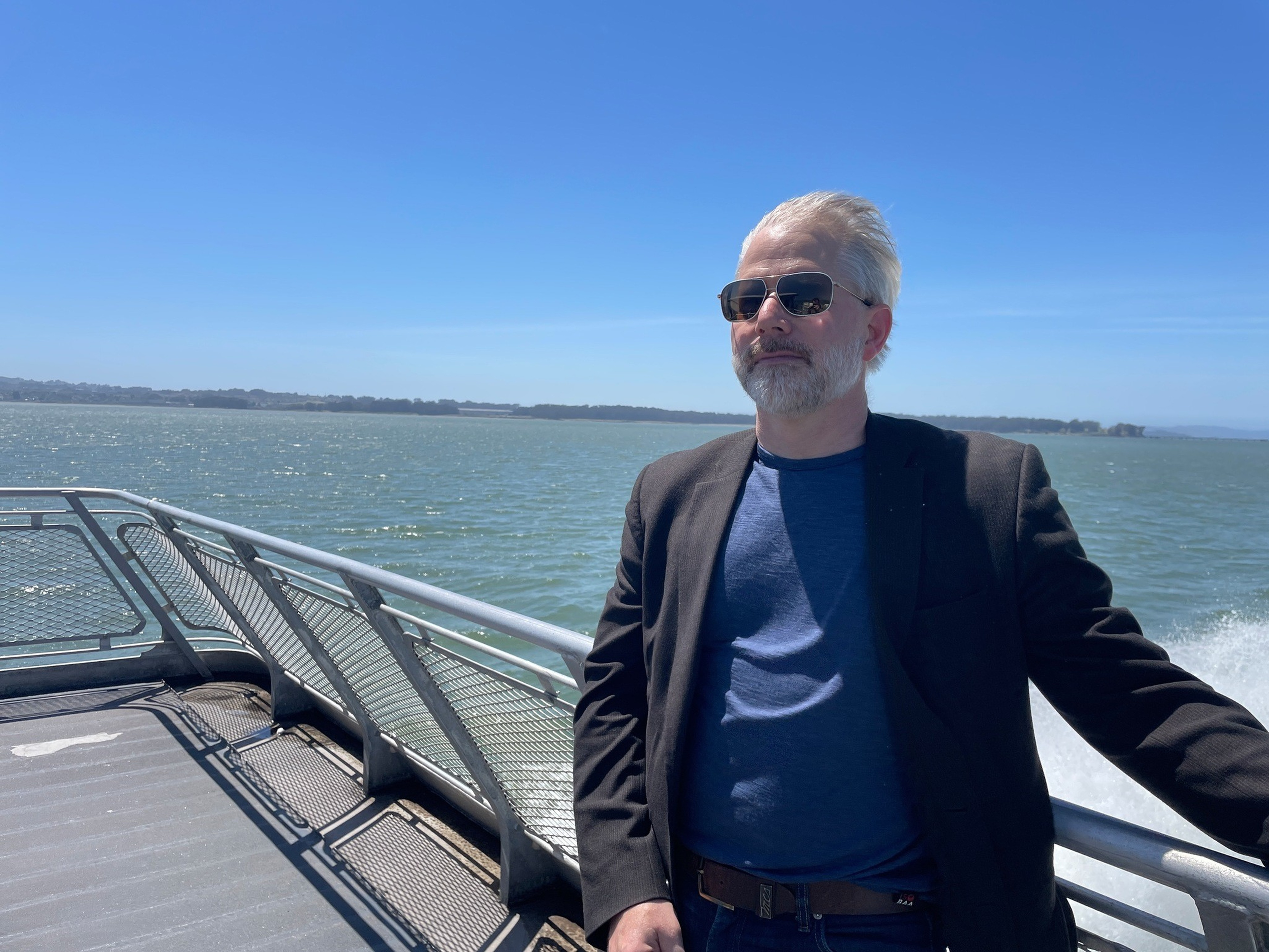 A man with white hair and a beard wears sunglasses, a blue shirt, and a dark blazer while standing on a boat with water and land in the background.