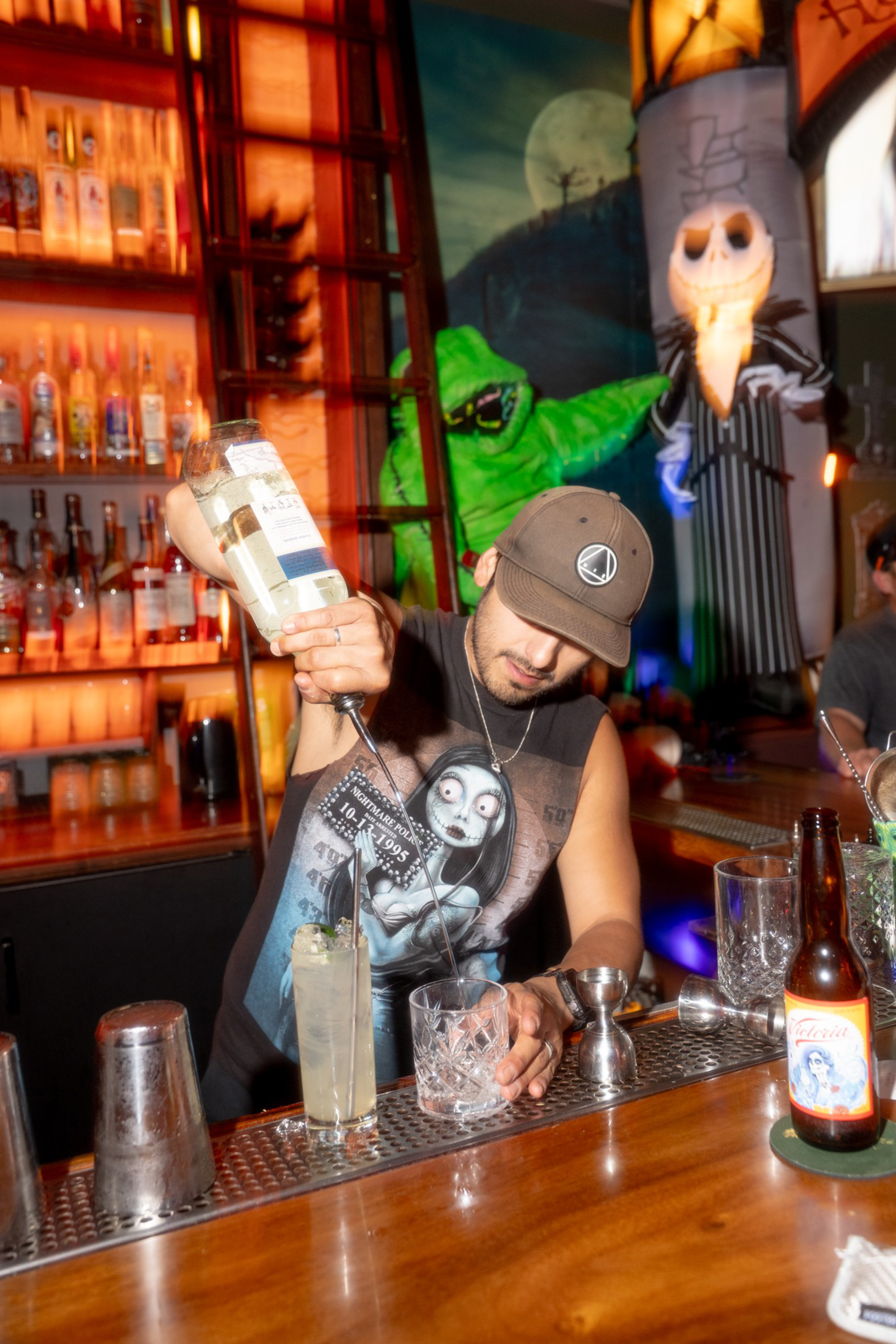 A bartender wearing a sleeveless shirt and cap pours liquor into a glass, with Halloween-themed decorations including green and skeleton figures behind him.