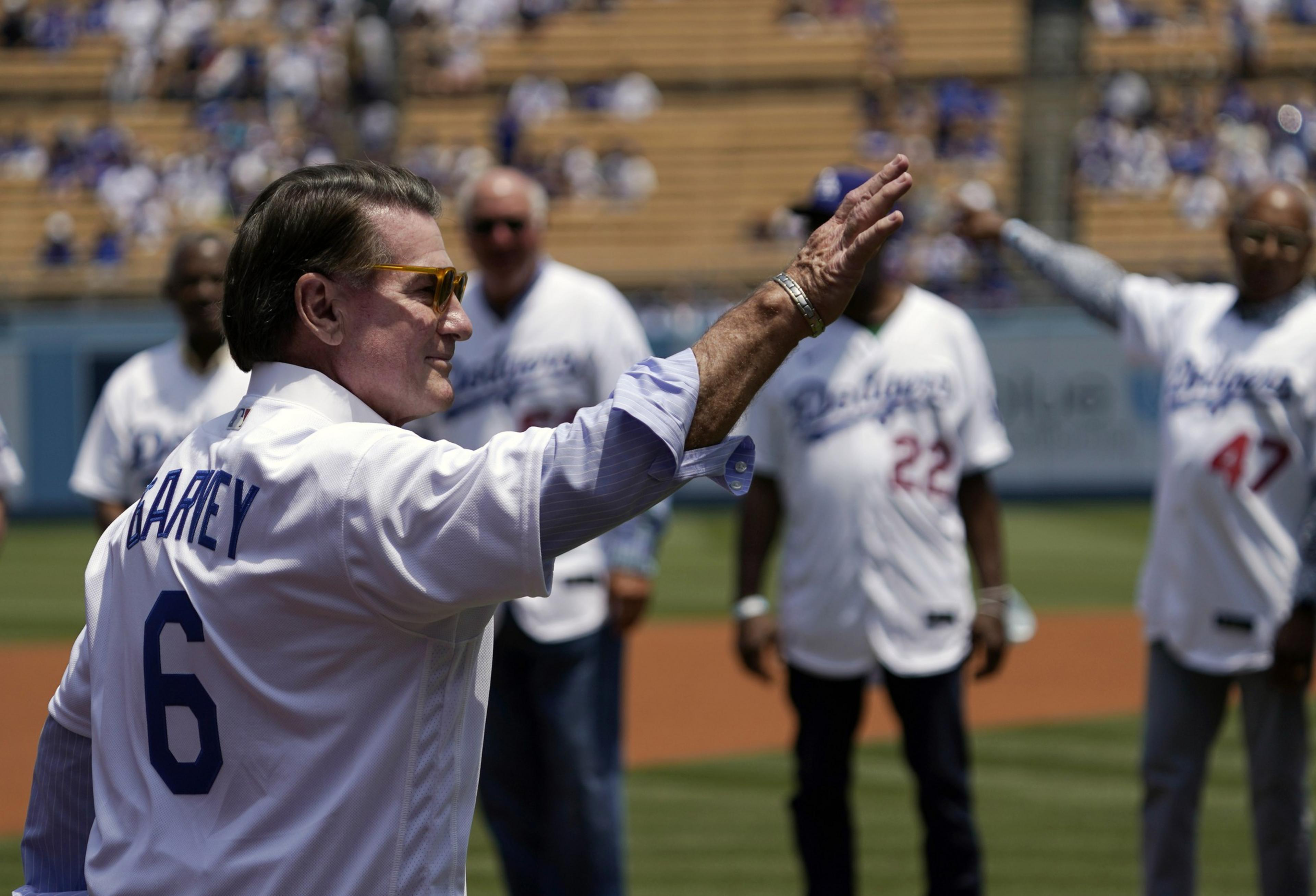 A man in sunglasses waves on a sunny baseball field, wearing a "Garvey 6" Dodgers jersey among other players.