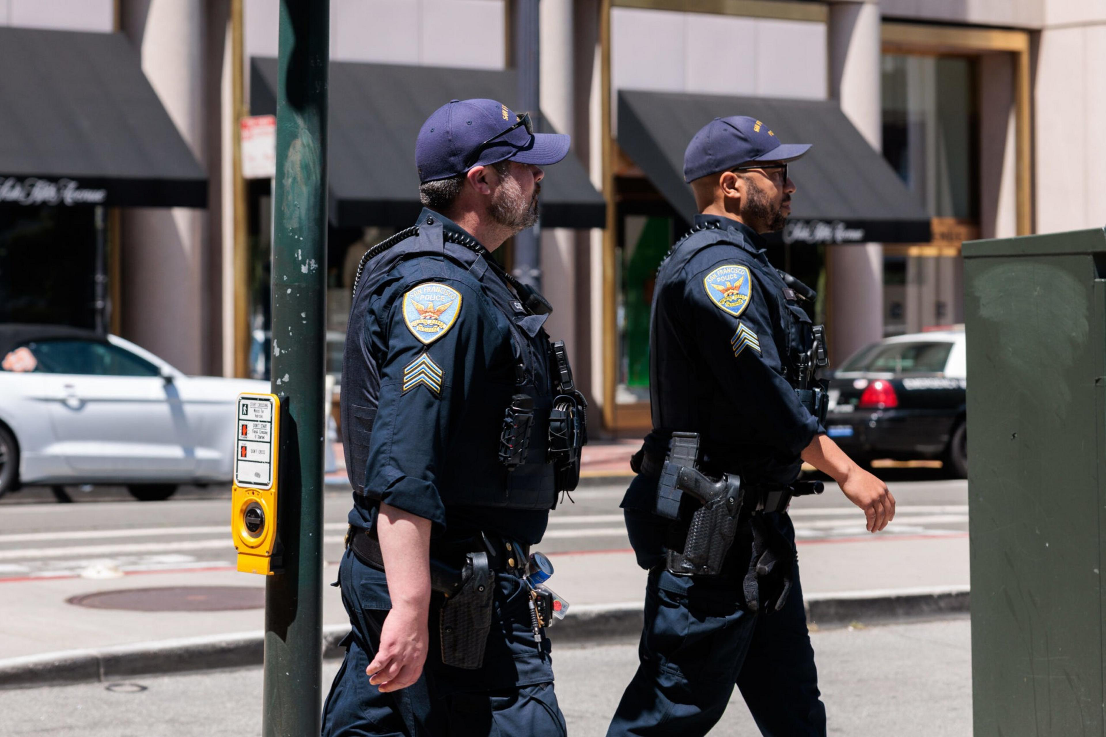 Two uniformed police officers in caps walk side by side on a city street near a pedestrian crossing button mounted on a pole.