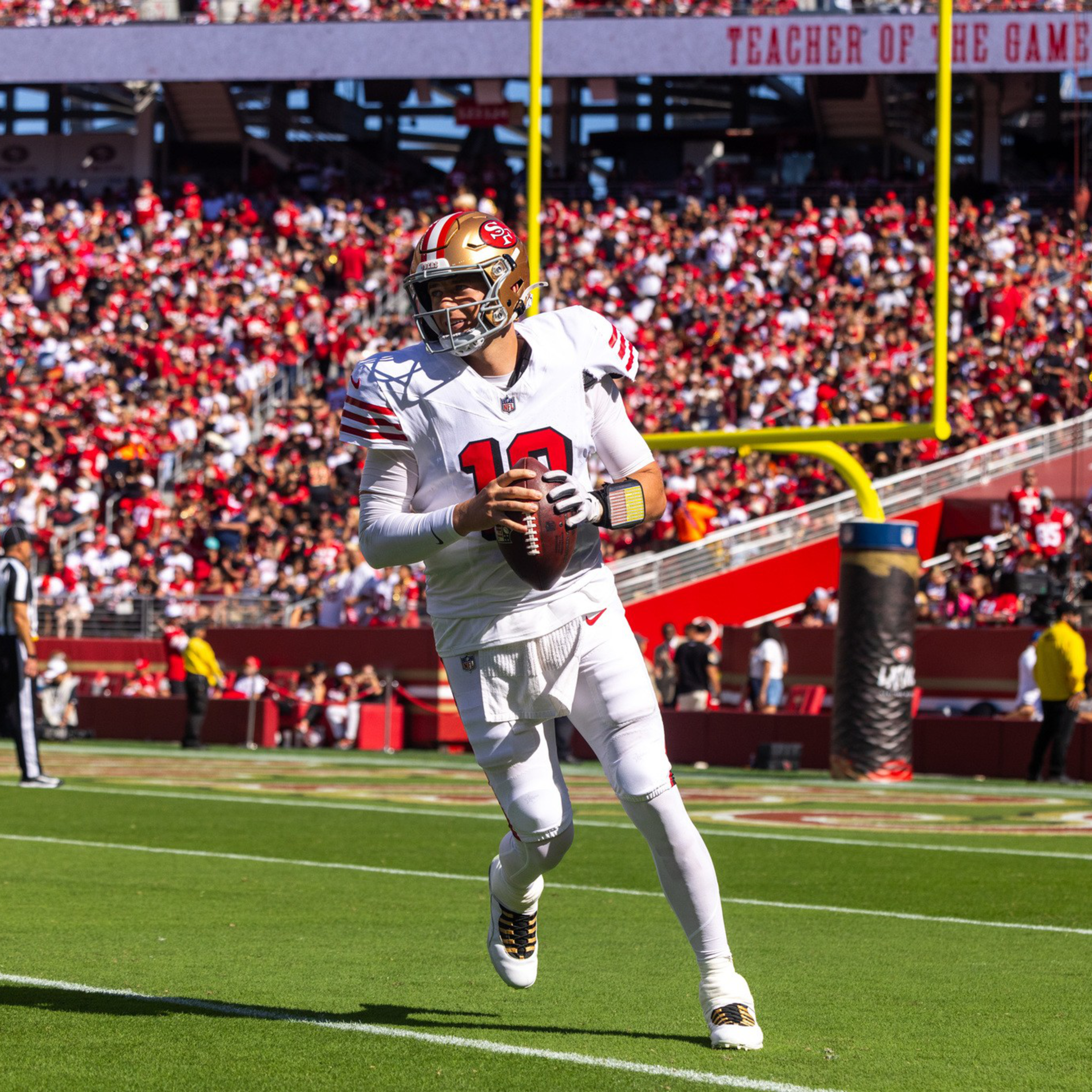 A football player in white and red gear with number 10 is holding the ball, running on a green field near the end zone with a crowd in the background.