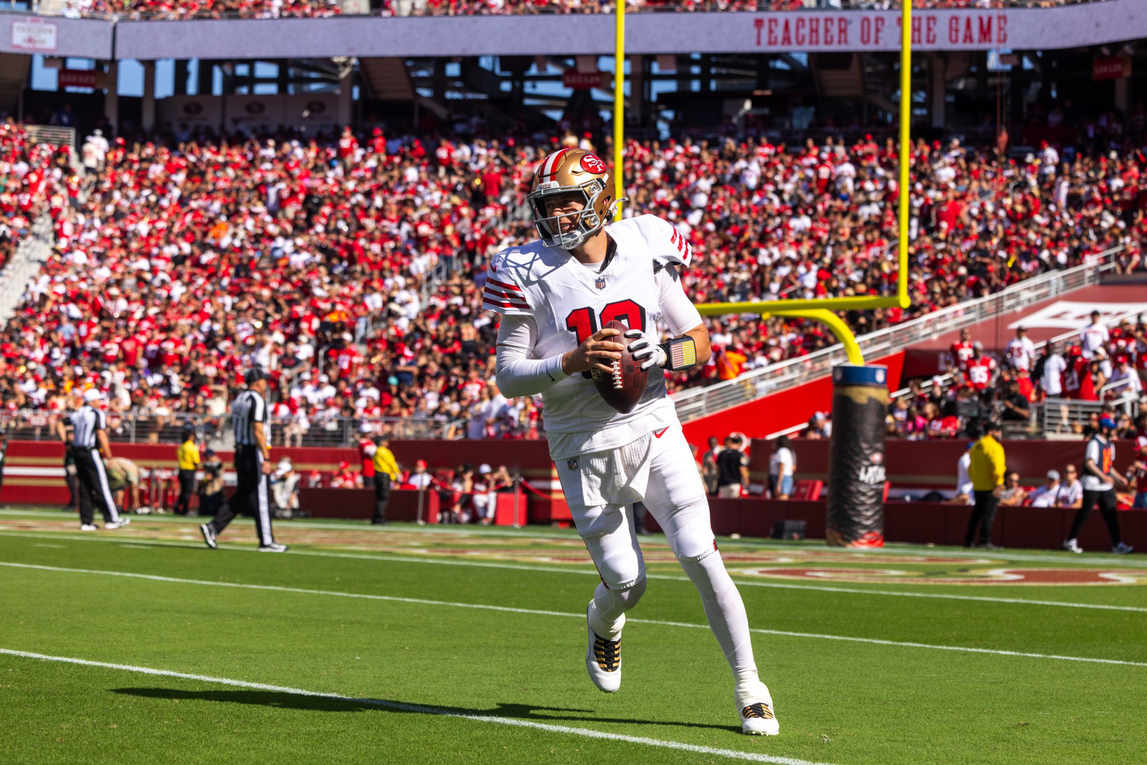 A football player in white and red gear with number 10 is holding the ball, running on a green field near the end zone with a crowd in the background.