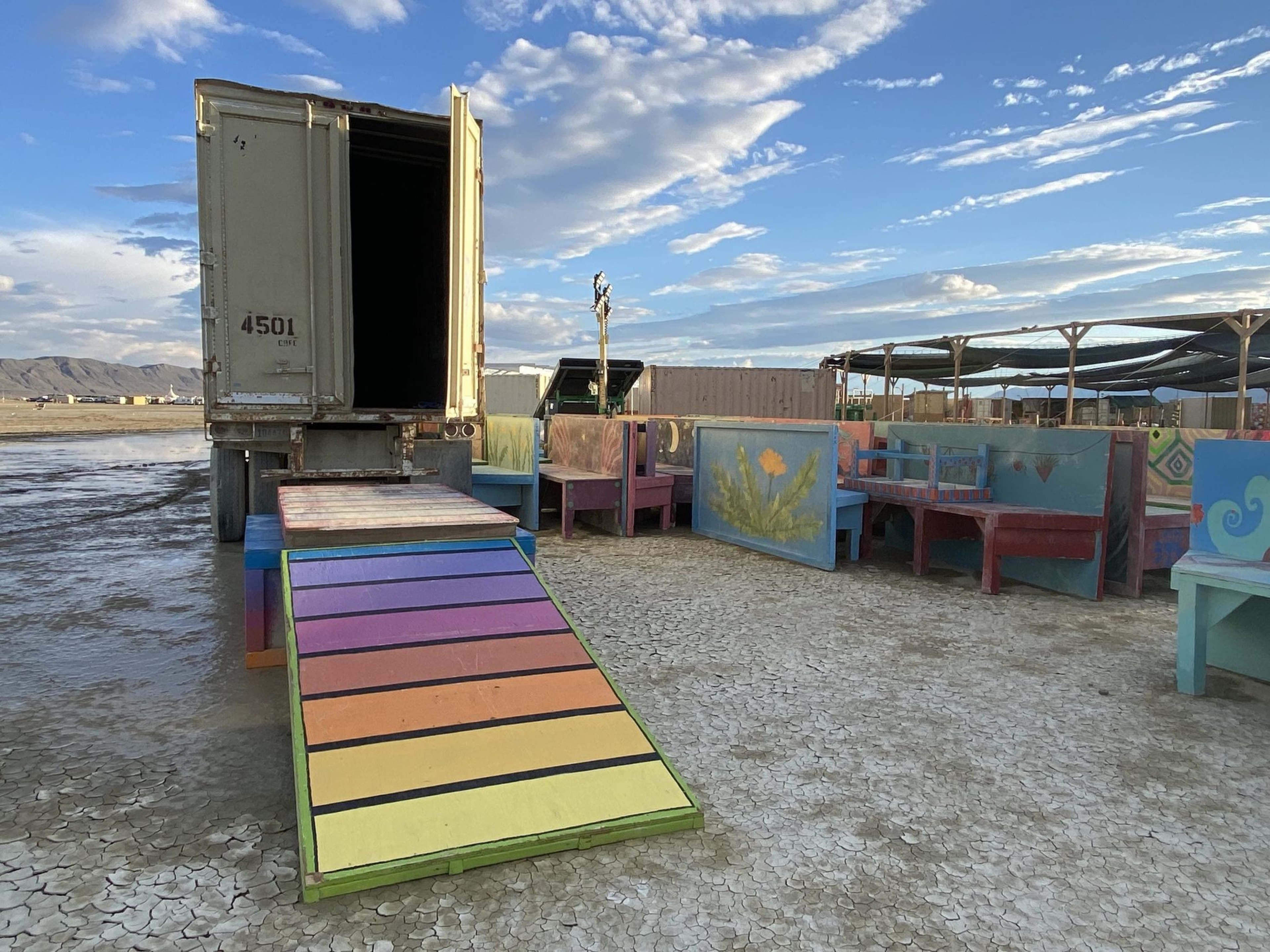 A colorful wooden ramp with rainbow stripes leads into an open truck trailer, surrounded by painted benches on dry, cracked ground under a blue sky.