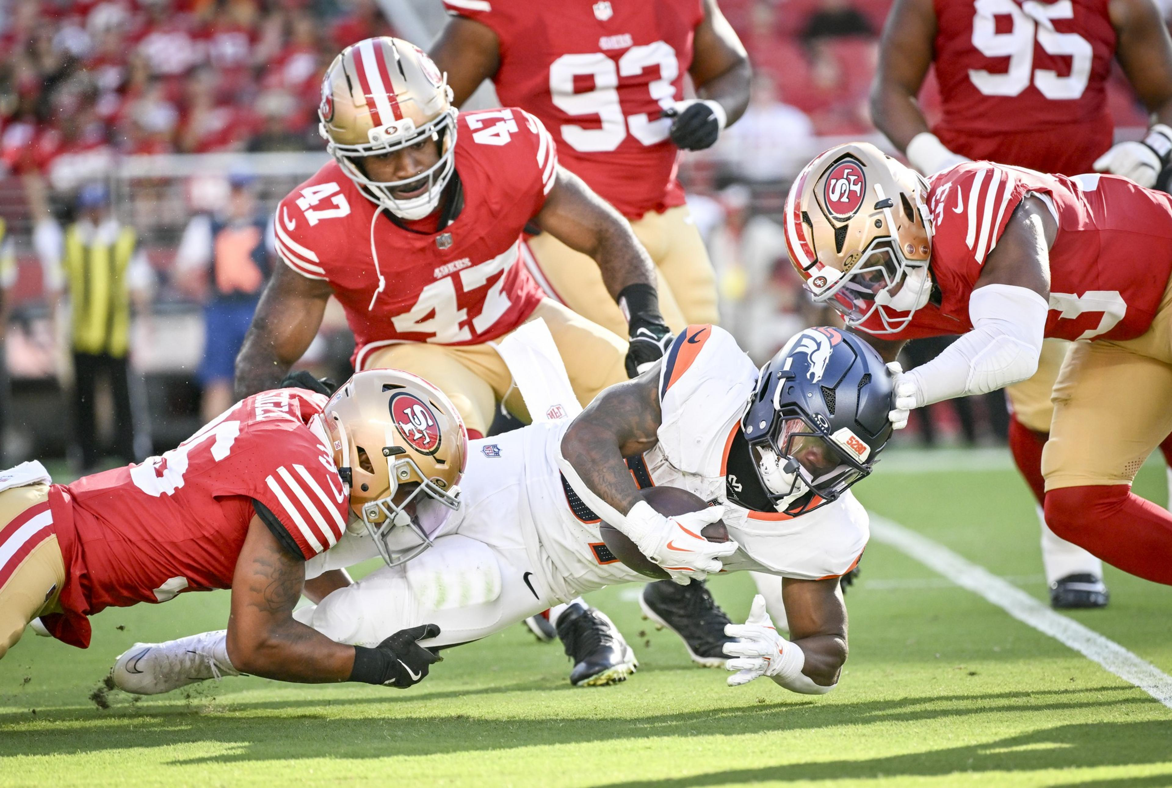 A football player in a white uniform is tackled by three opponents in red and gold uniforms on a green field.