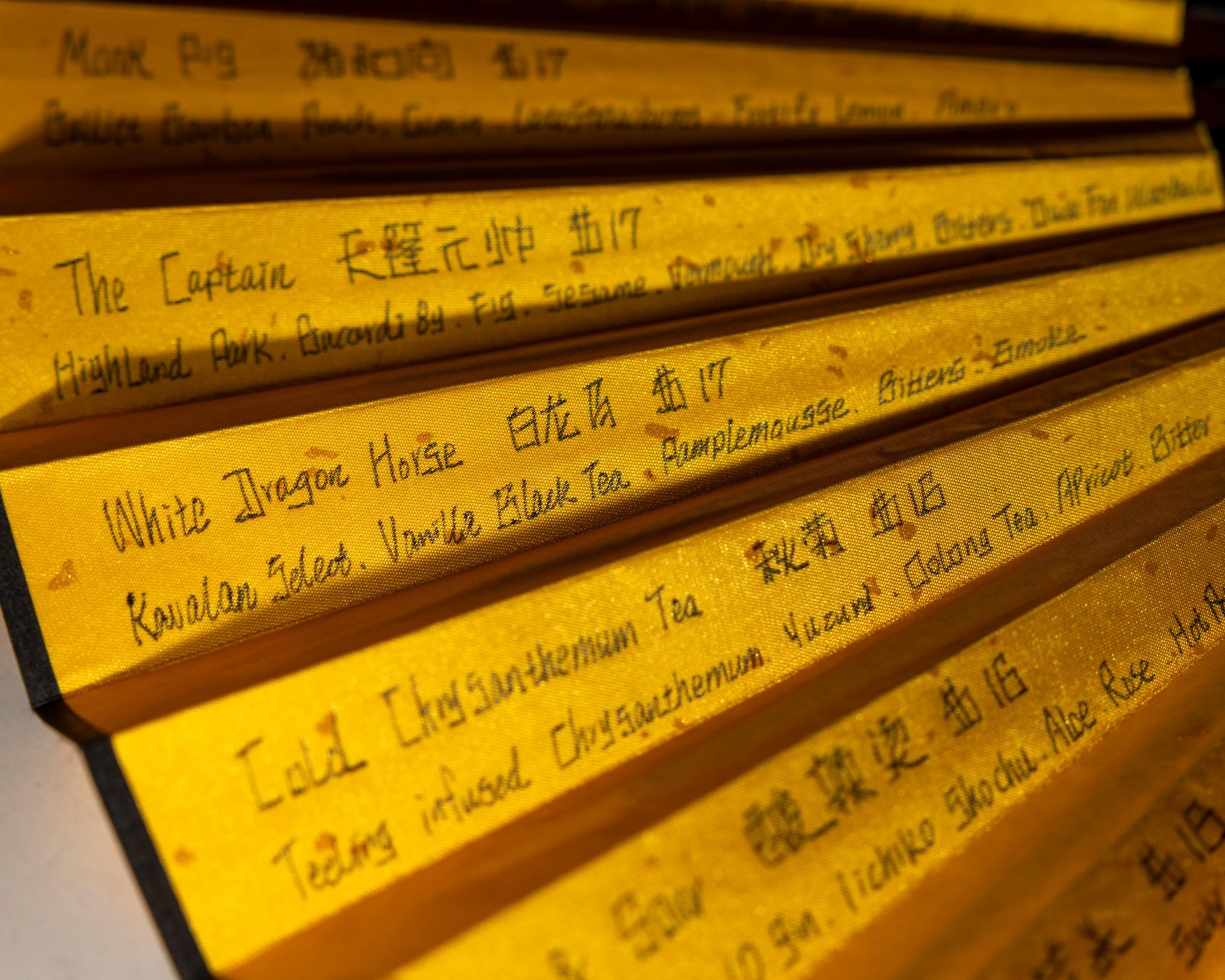 The image shows sections of a golden-colored folding hand fan with handwritten text in English and Chinese, listing various drink names and ingredients.
