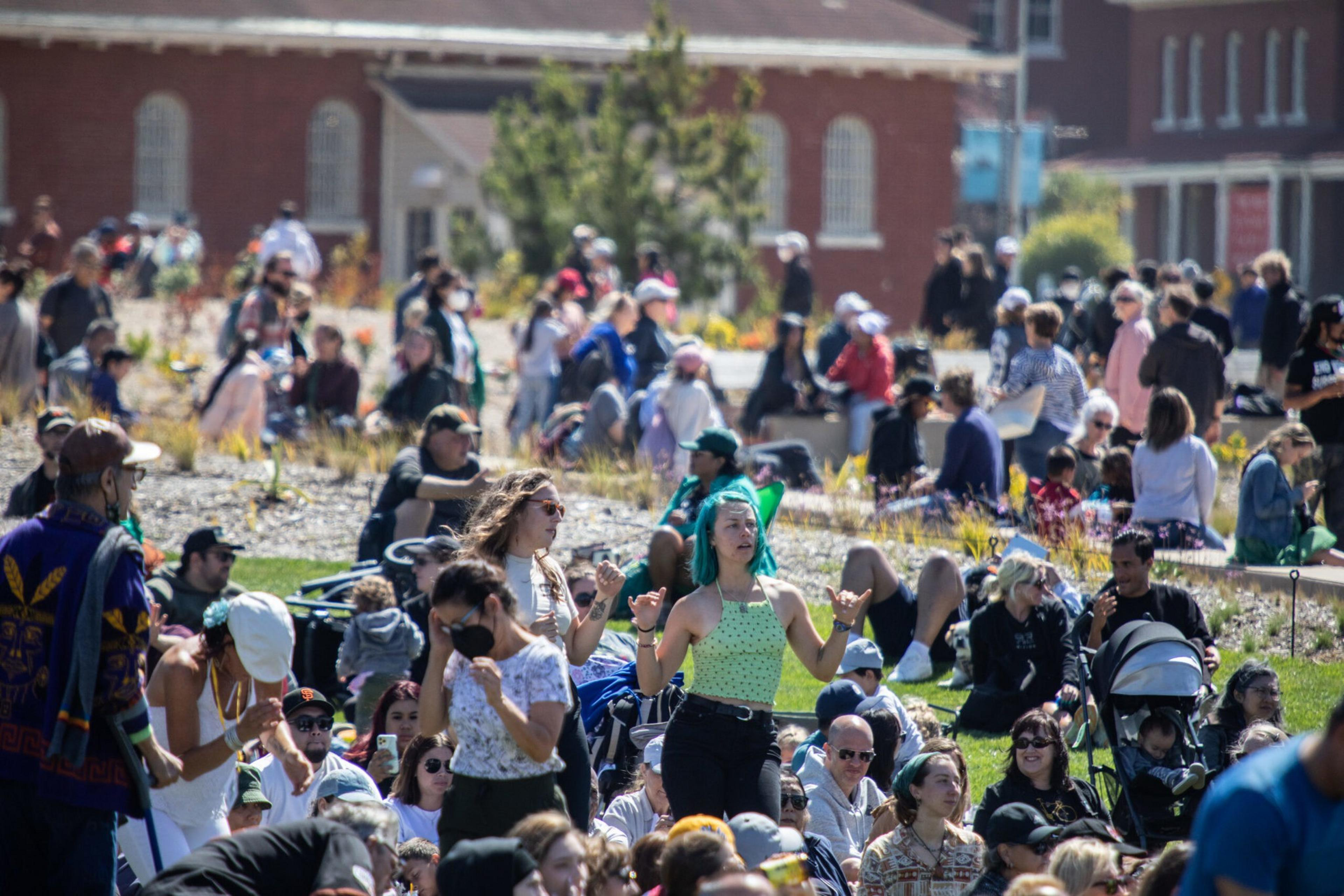 A large crowd of people gathers outdoors on grass and gravel near buildings, with some standing, sitting, or walking, engaging in various activities on a sunny day.