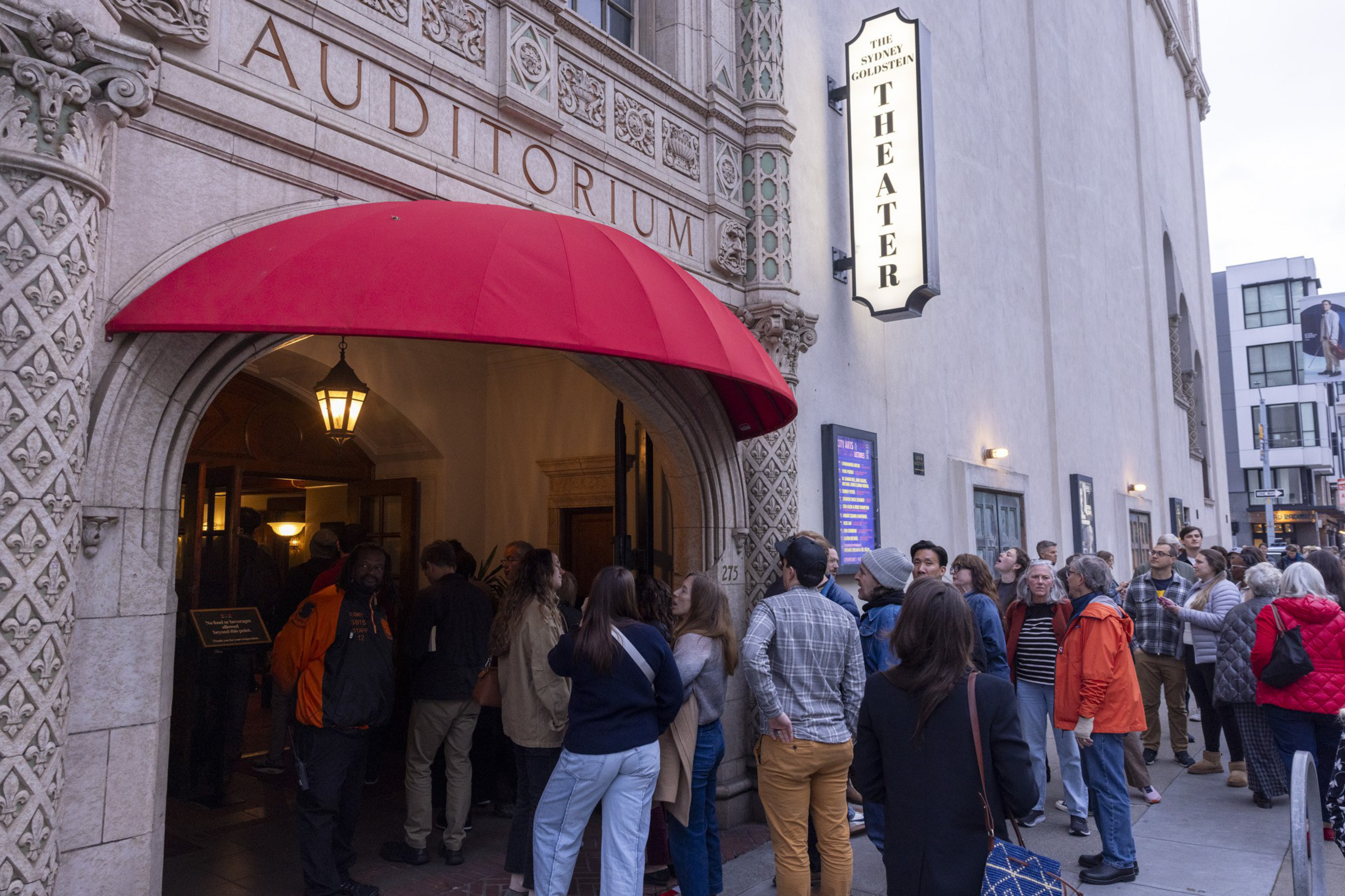 A crowd waits outside the Sydney Goldstein Theater, beneath a red awning, with decorative architecture, while a security guard stands by the entrance.