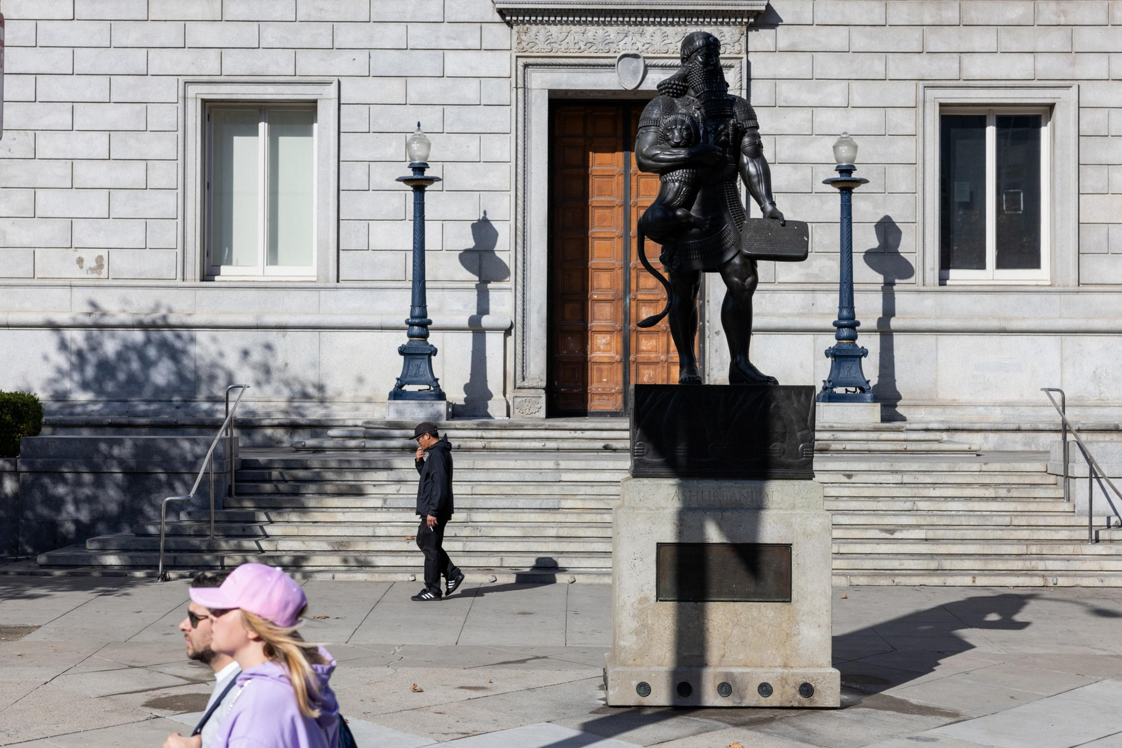 People walk through an empty courtyard area that has a large statue of a standing bearded man on a pedestal.