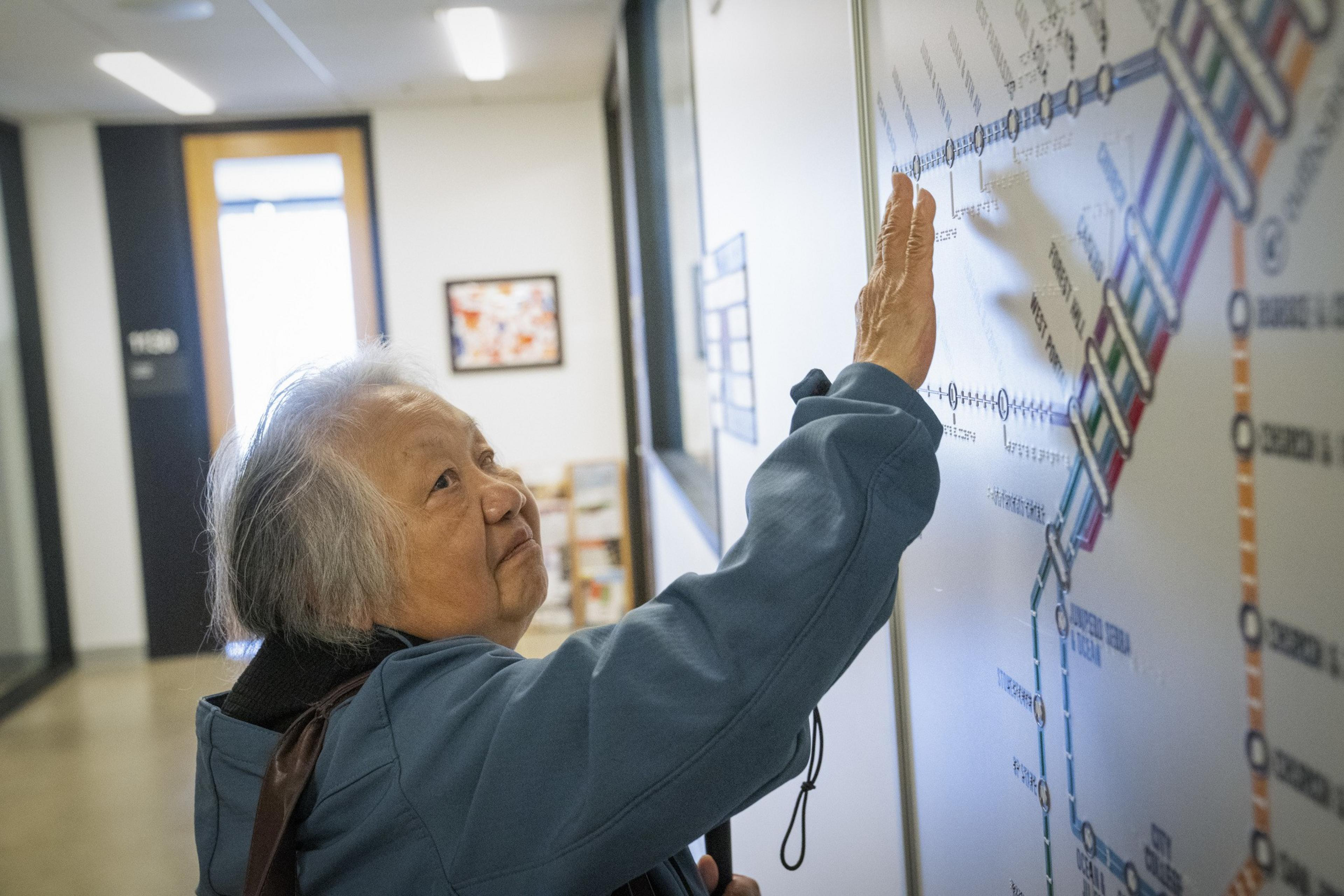 An elderly person in a blue jacket is touching a detailed transit map on a wall, seemingly studying it closely. The room is well-lit with a doorway and art in the background.
