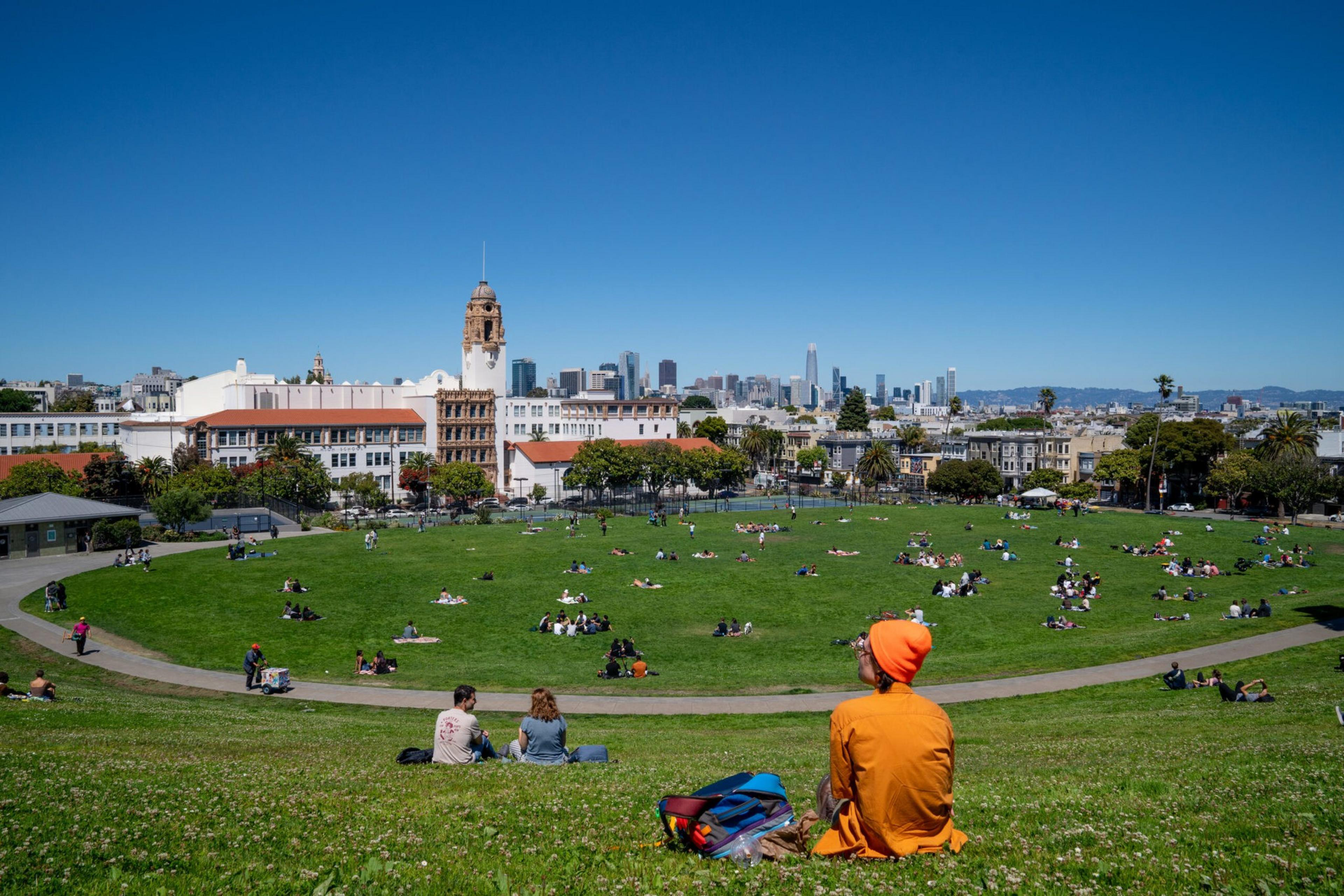 A large circular park with many people sitting and relaxing on the grass, surrounded by historic buildings and a distant city skyline under a clear blue sky.