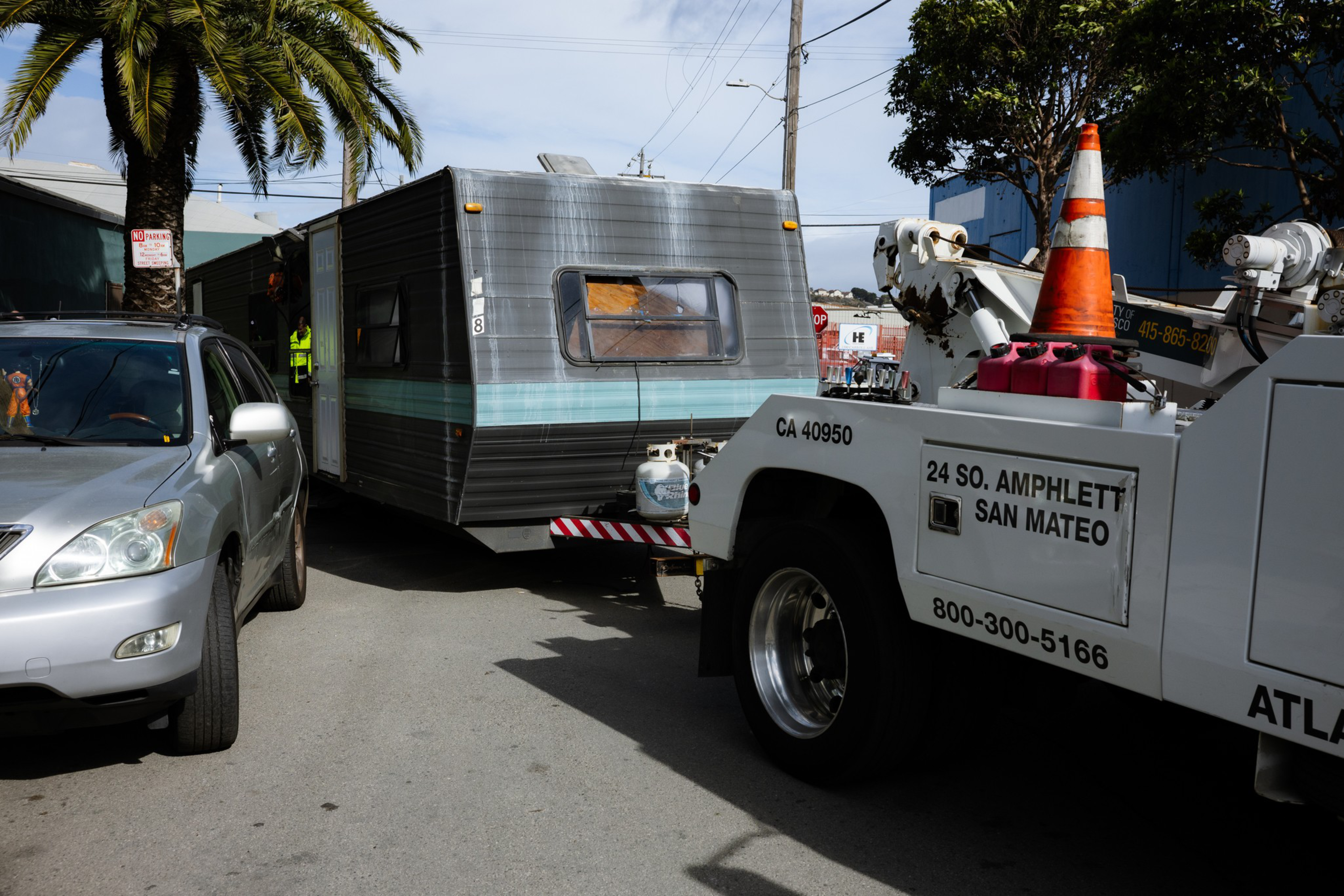 A tow truck is towing a gray and teal camper trailer off a street with a silver SUV parked nearby and a palm tree in the background.