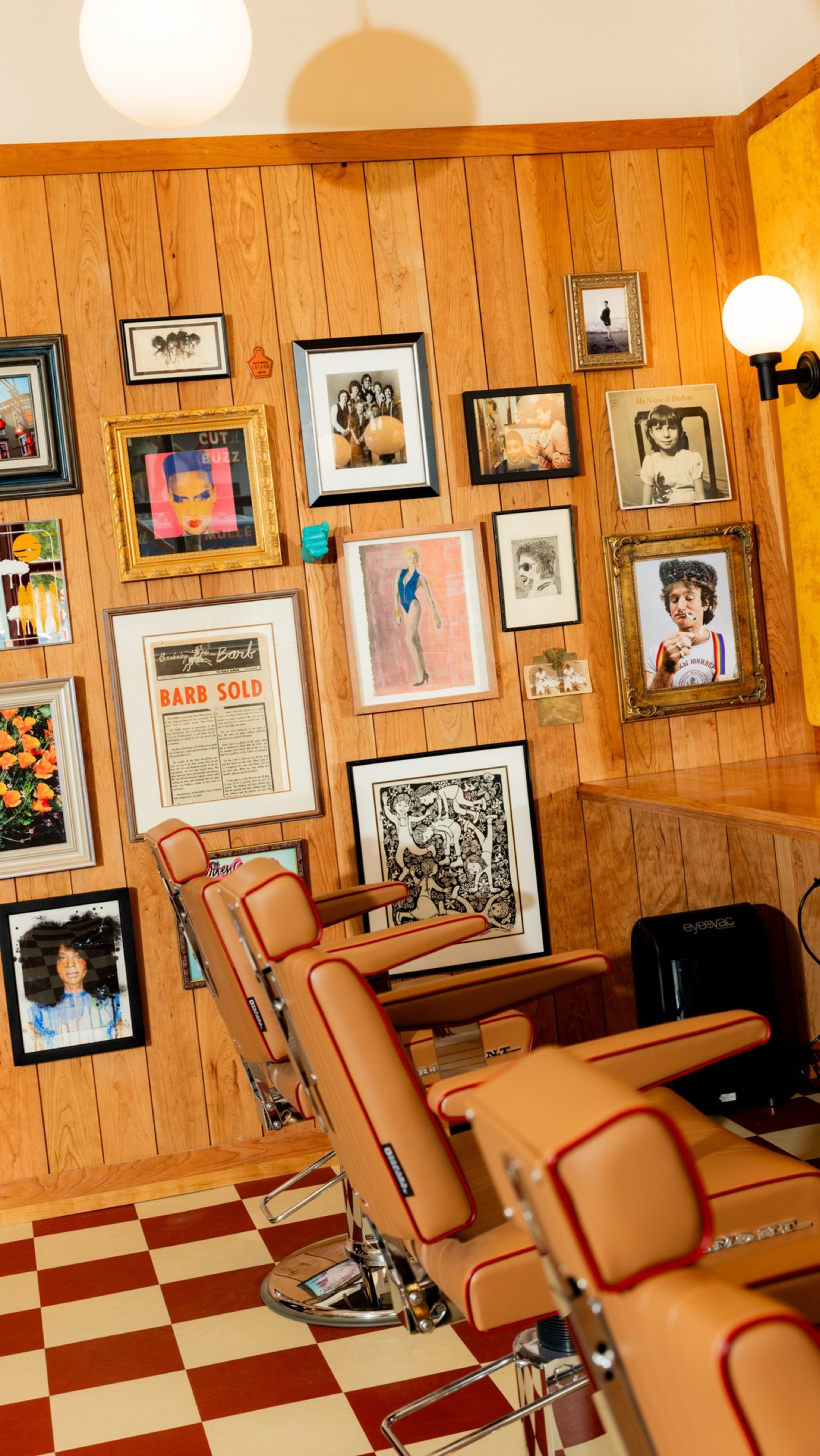 Three vintage-style barber chairs with orange upholstery sit in front of a wood-paneled wall covered in various framed photos and artwork.
