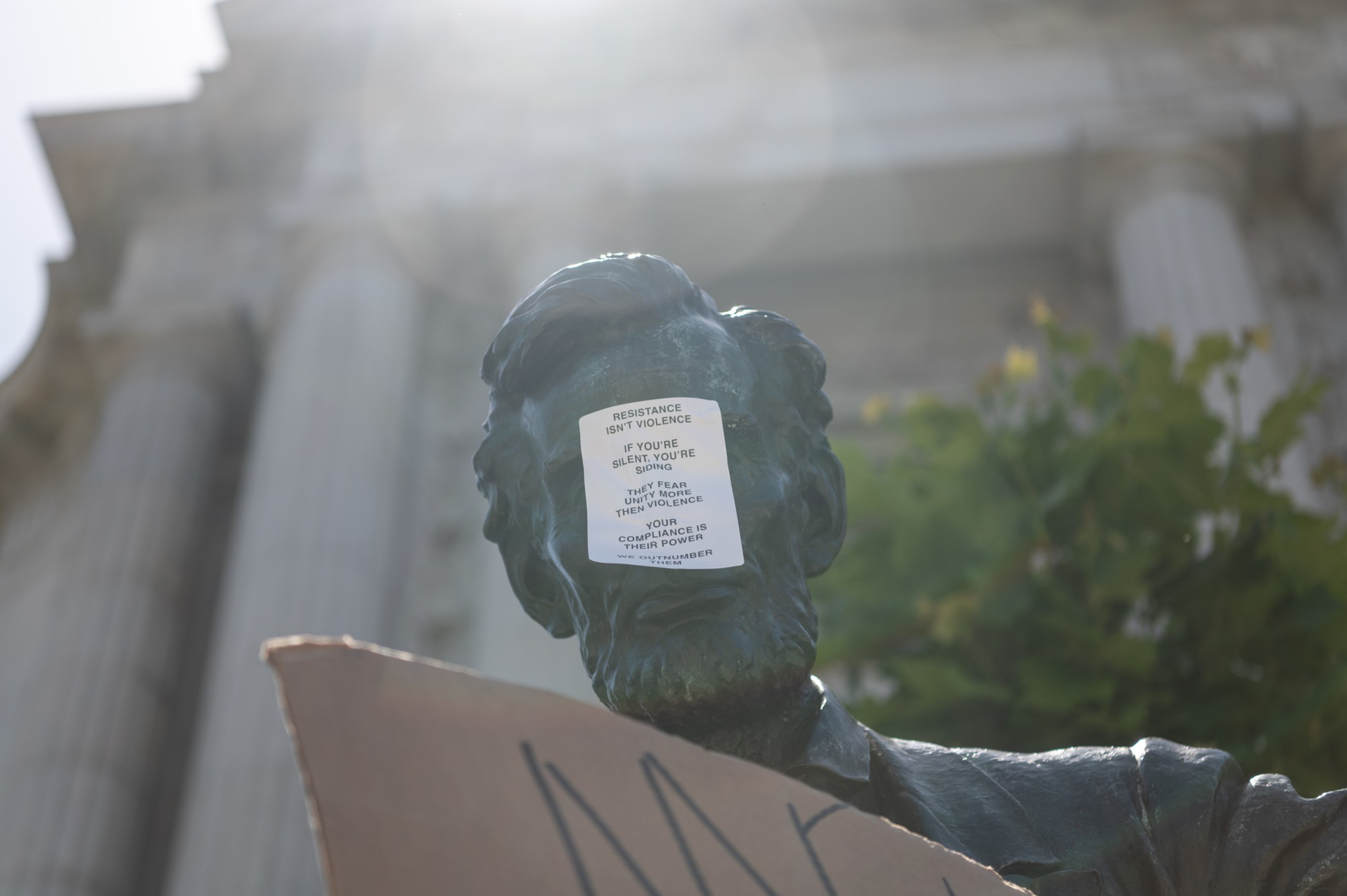 A statue has a sticker on its face that reads, &quot;Resistance isn't violence. If you're silent, you're siding. They fear unity more than violence. Your compliance is their power.&quot;
