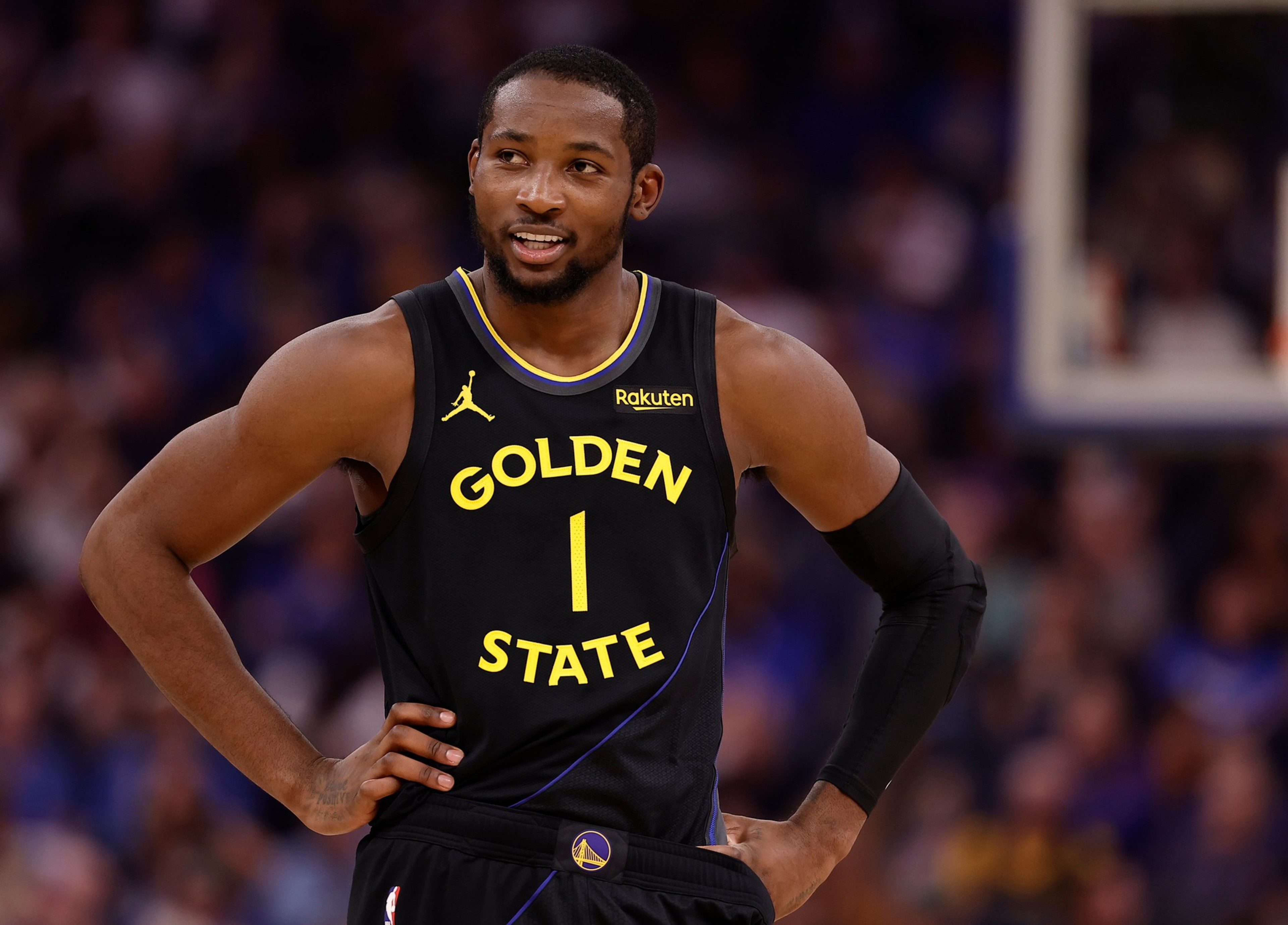A basketball player in a black Golden State Warriors jersey with the number 1 stands with hands on hips, smiling during a game.