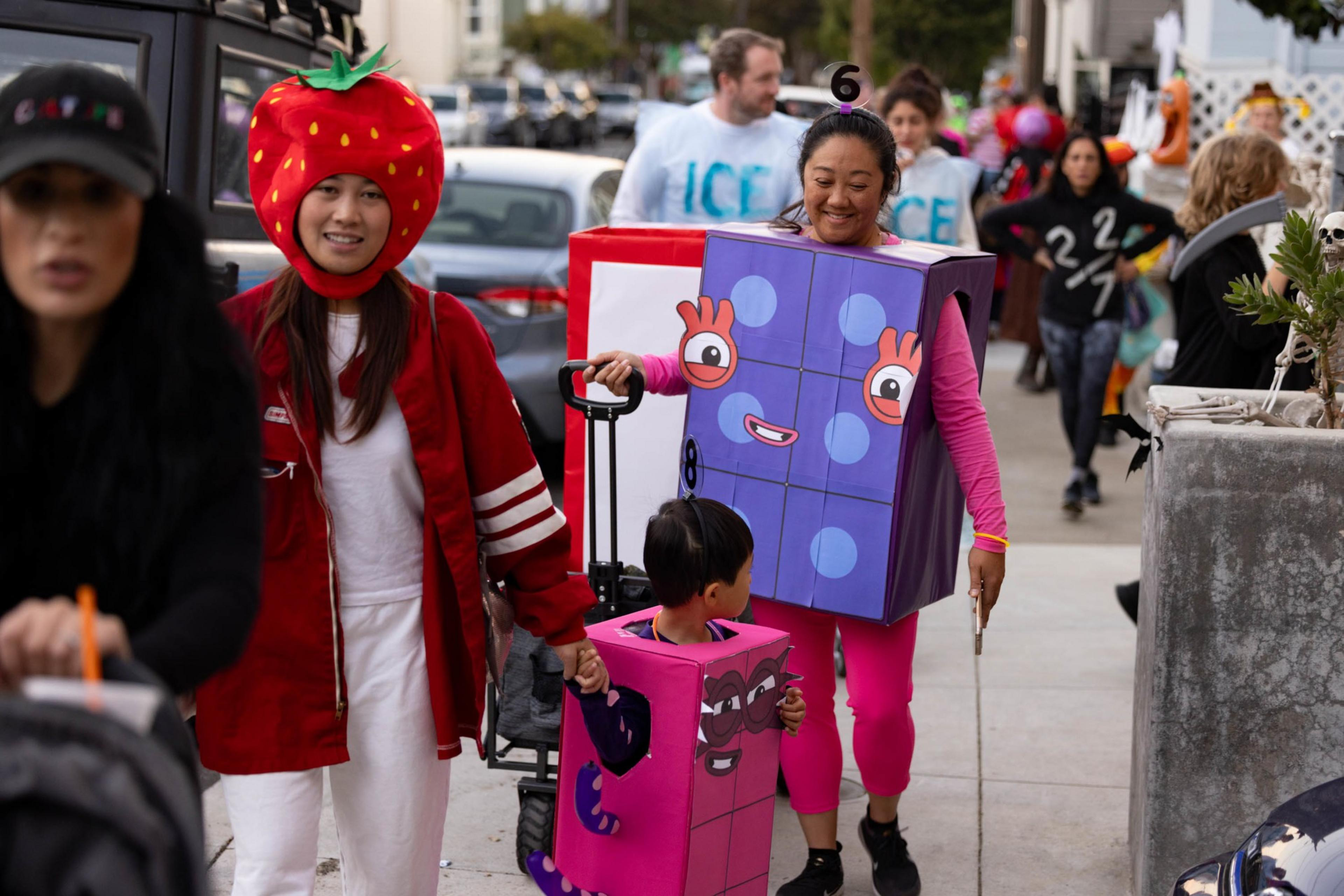 A woman and child wear colorful Tetris block costumes while another woman beside them wears a large strawberry hat, all walking on a busy sidewalk.