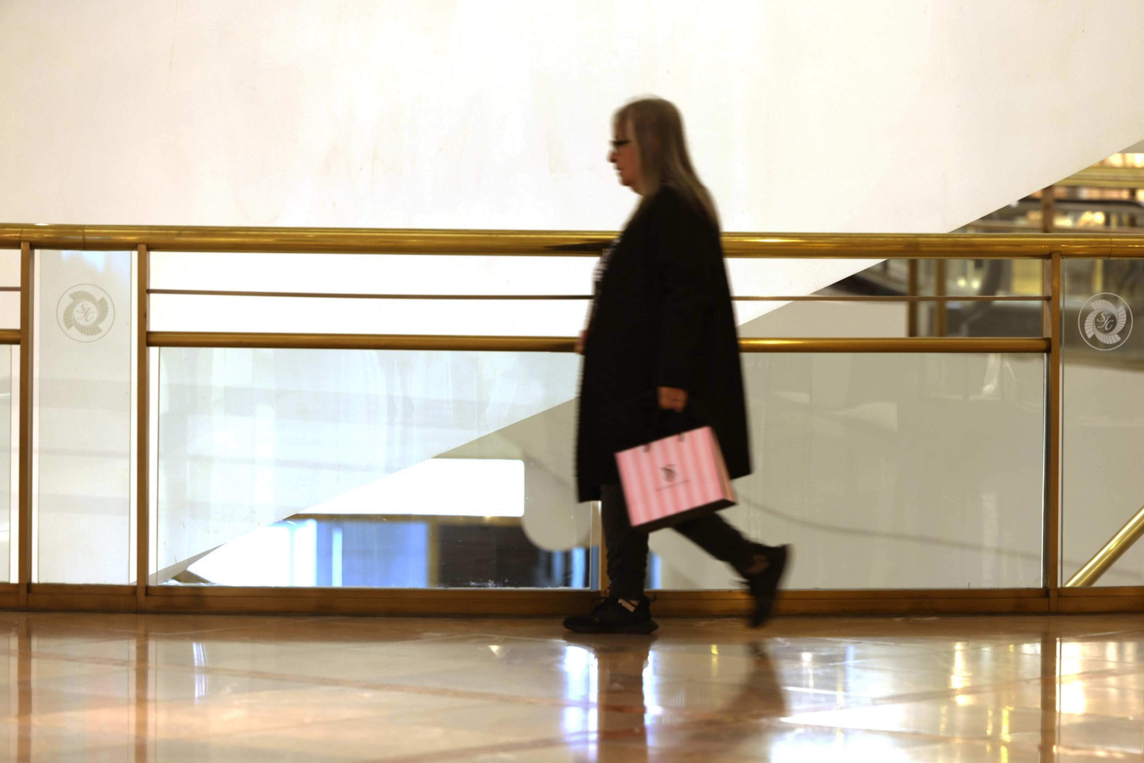 A person in long attire holding a pink bag walks past a brass-railed, light-filled interior.