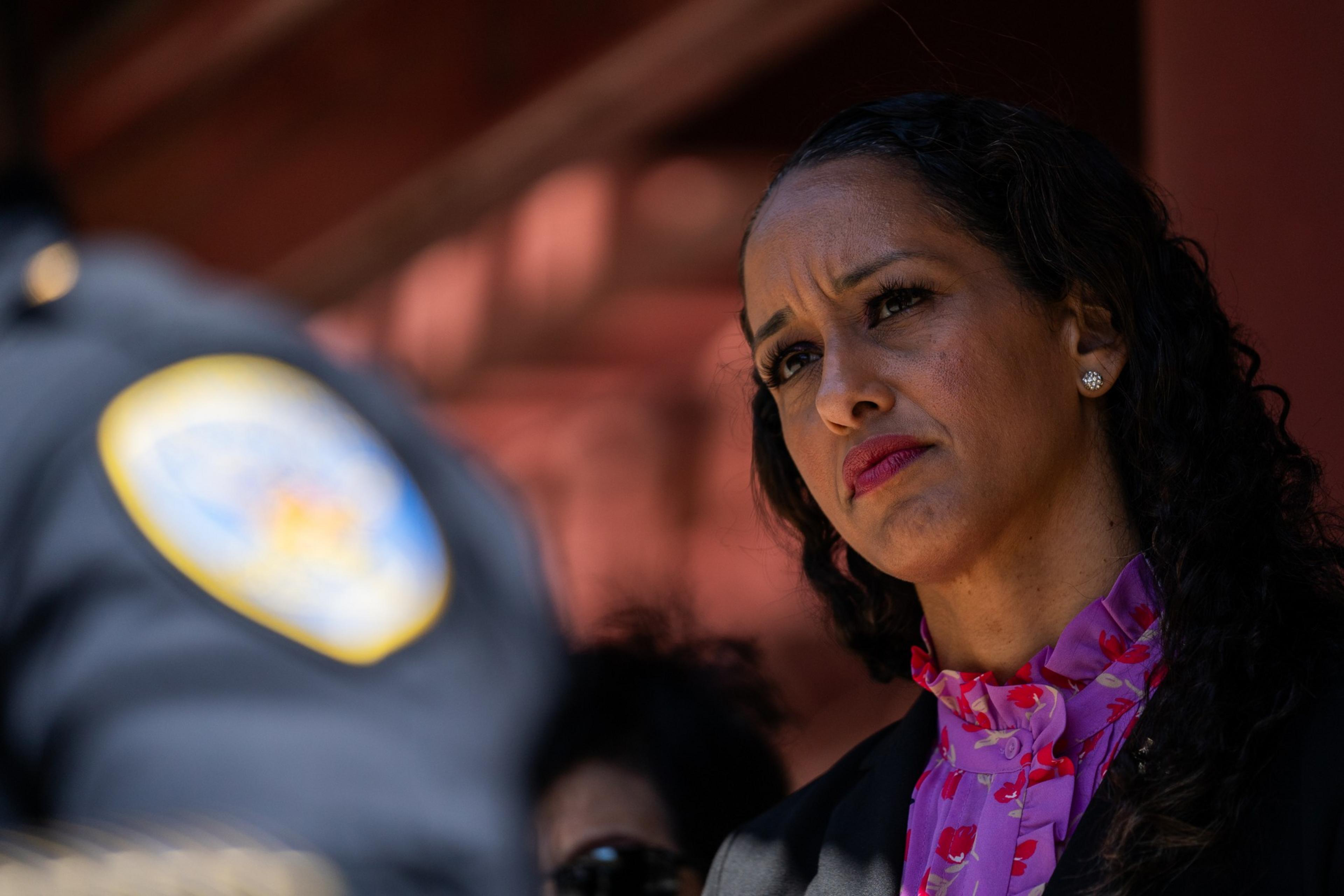 A woman wearing a purple blouse looks into the distance as a portion of a police uniform is in the foreground.