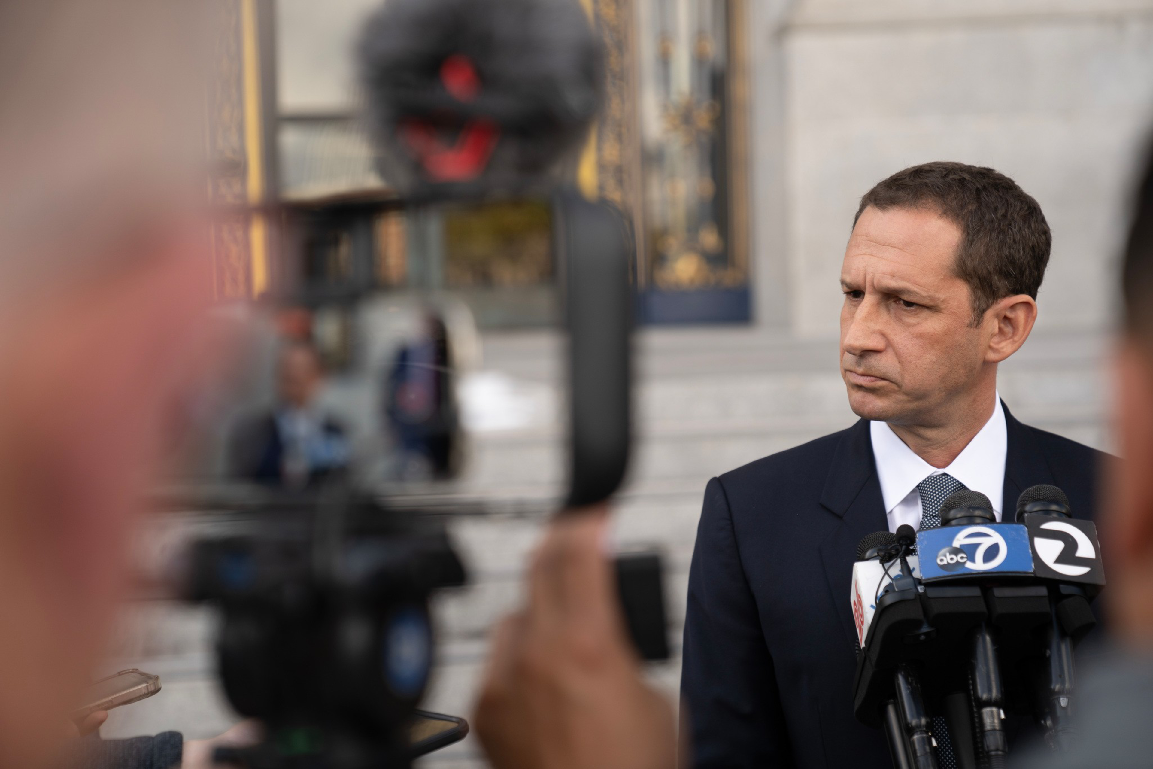 A man in a dark suit and white shirt stands in front of microphones from news outlets, looking serious, with a blurred crowd and building in the background.