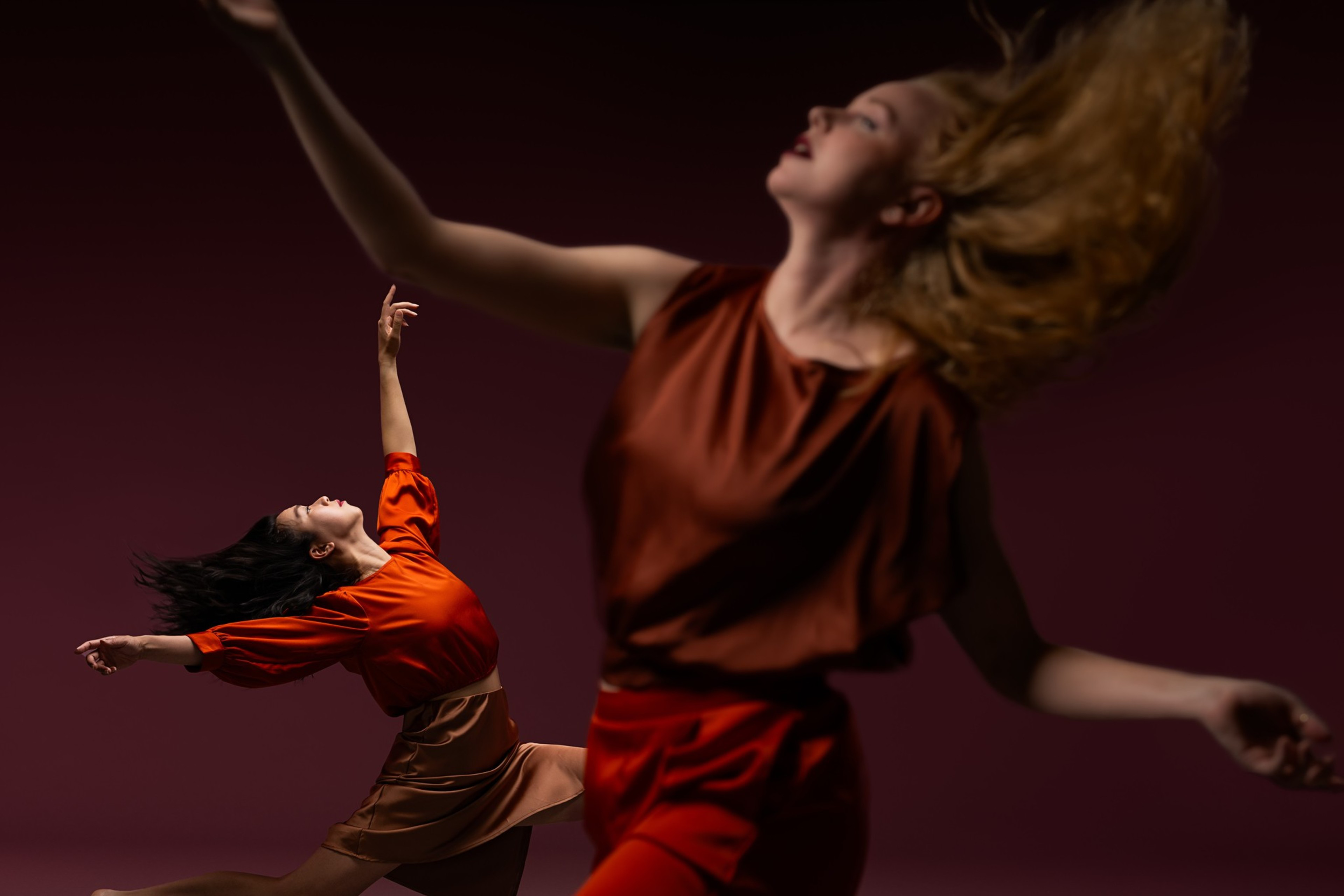 Two dancers in motion against a deep red background. One wears a red top and brown skirt, with arms gracefully extended. The other's image is blurred in the foreground.