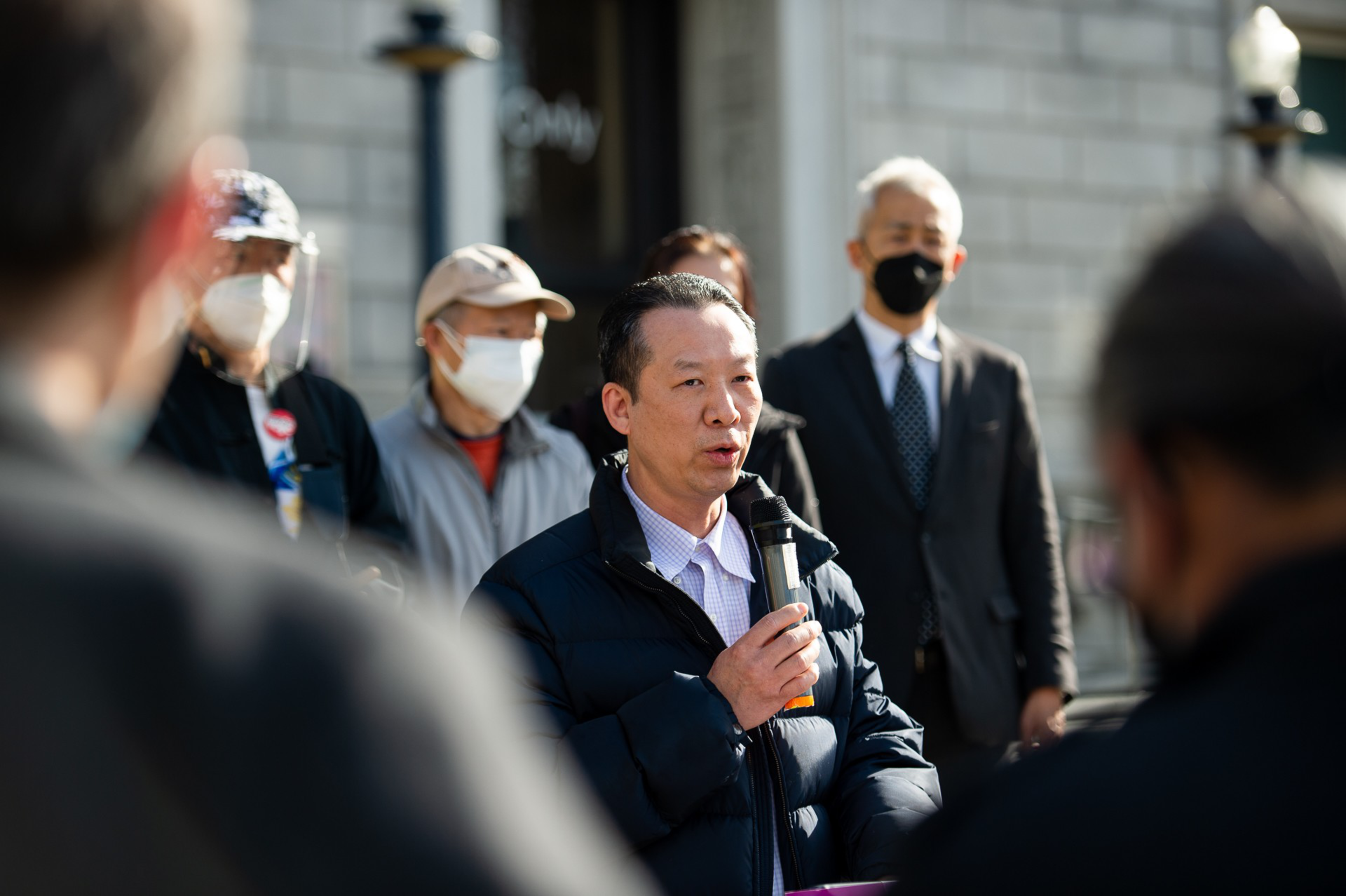 A man in a dark jacket speaks into a microphone while several people, some wearing masks, stand behind him outside a building.