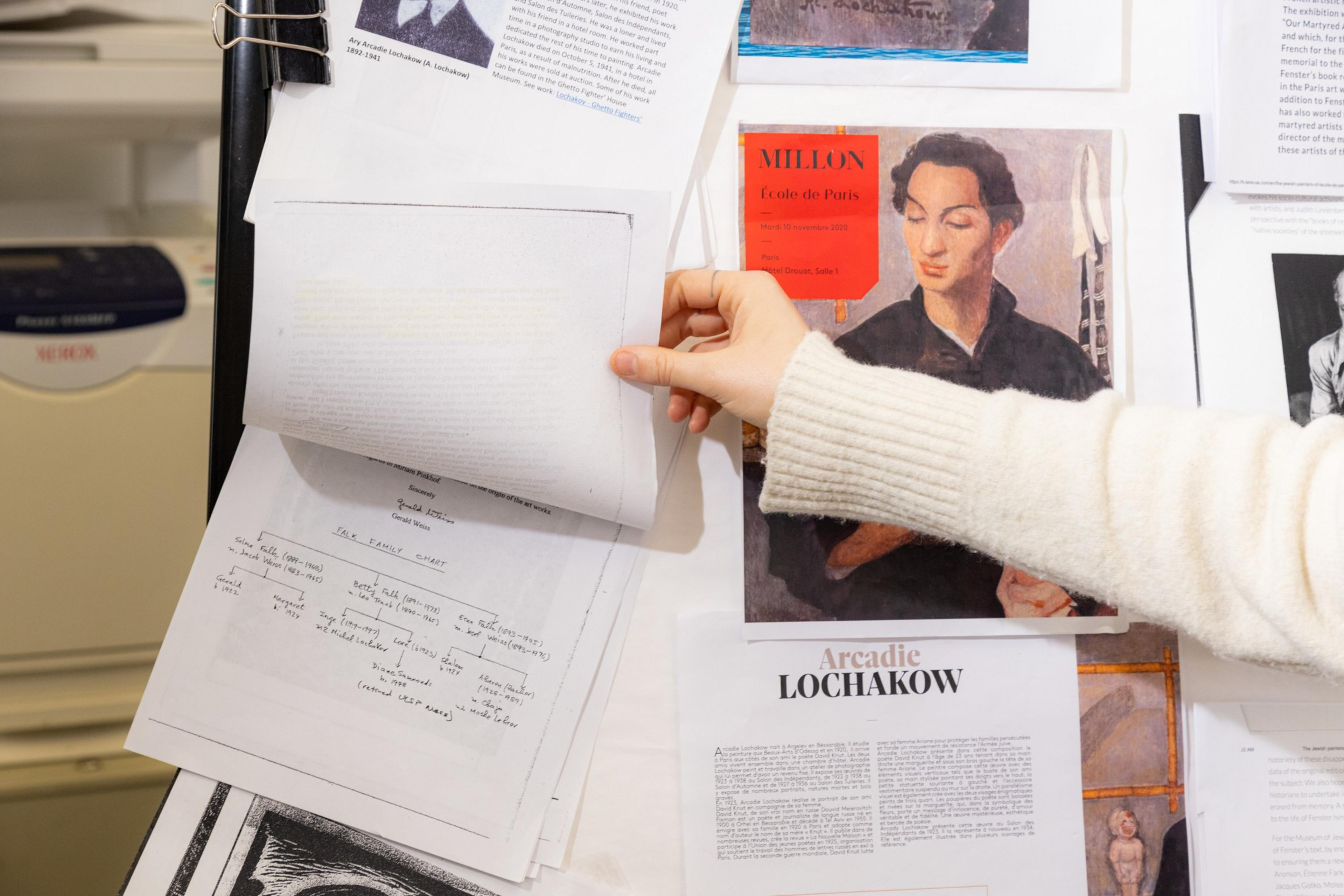 A hand flips through papers on a bulletin board with various articles, one featuring a painted portrait of a person.