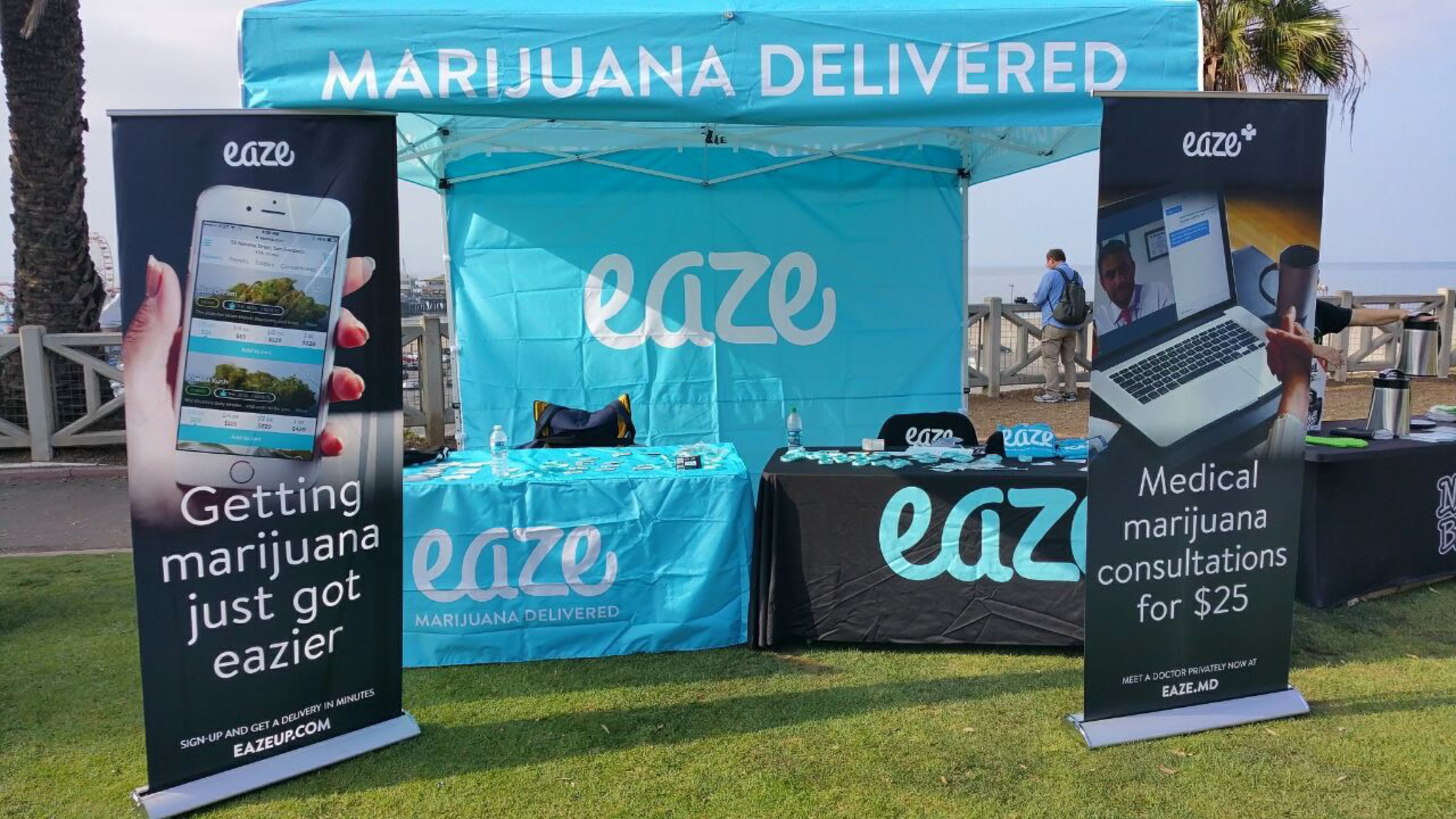 A booth with blue and black &quot;Eaze&quot; banners, advertising marijuana delivery and consultations. It features promotional materials on tables and large display banners.