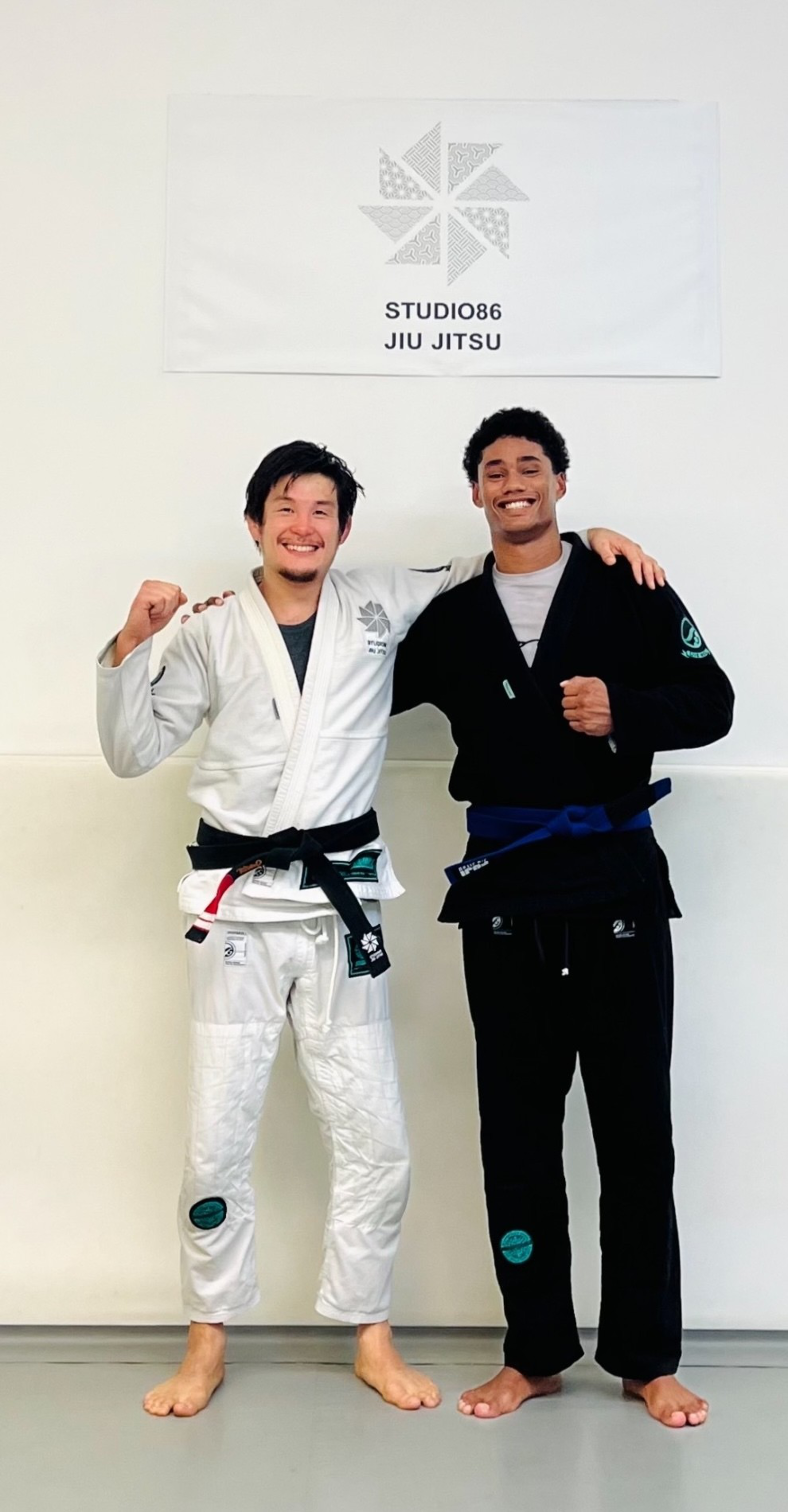 Two men stand barefoot on a mat, smiling and posing in martial arts gis; one wears a white gi with a black belt, the other a black gi with a blue belt.