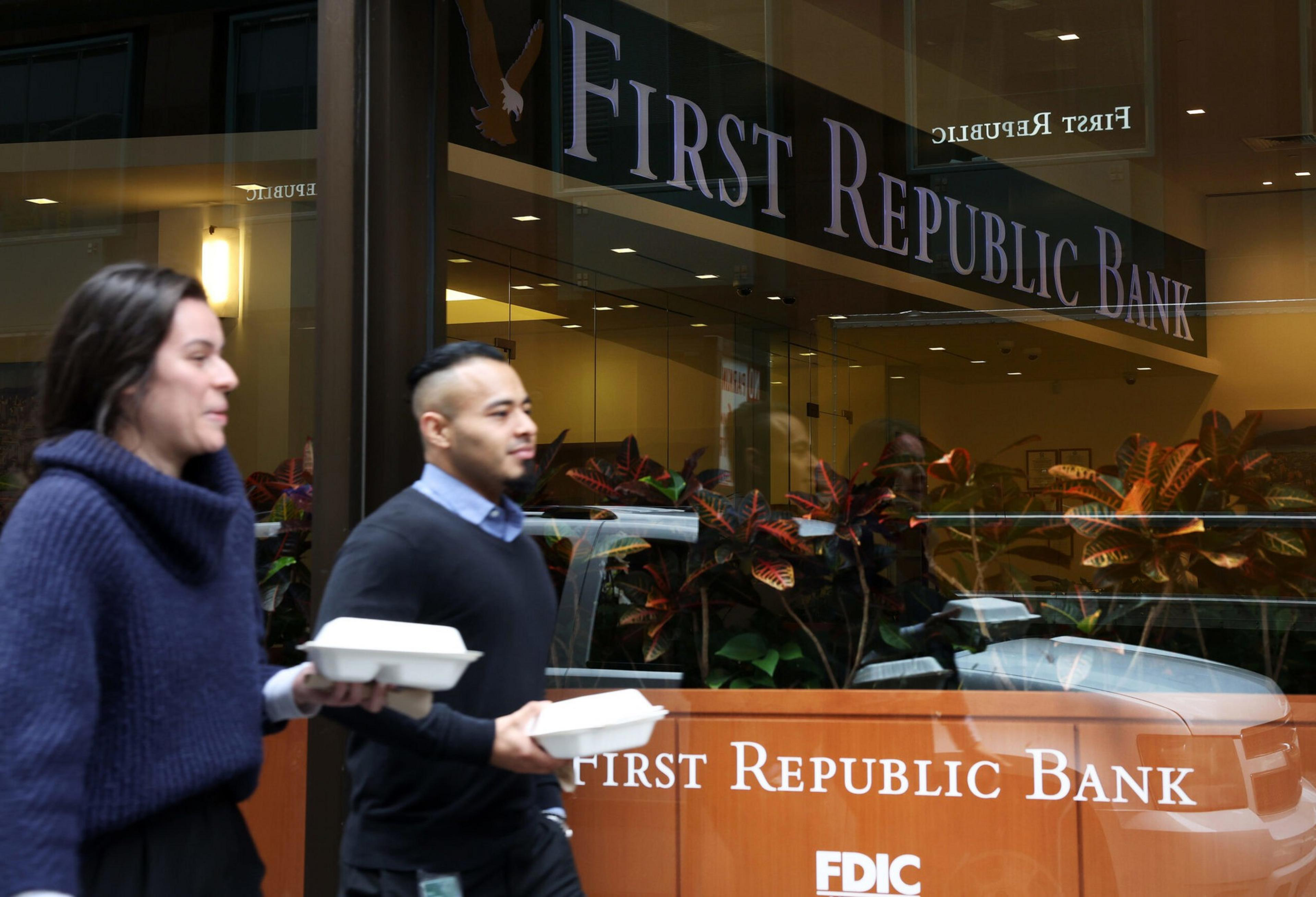 Two people walk past a First Republic Bank branch, carrying food containers. The bank's sign and FDIC logo are visible.