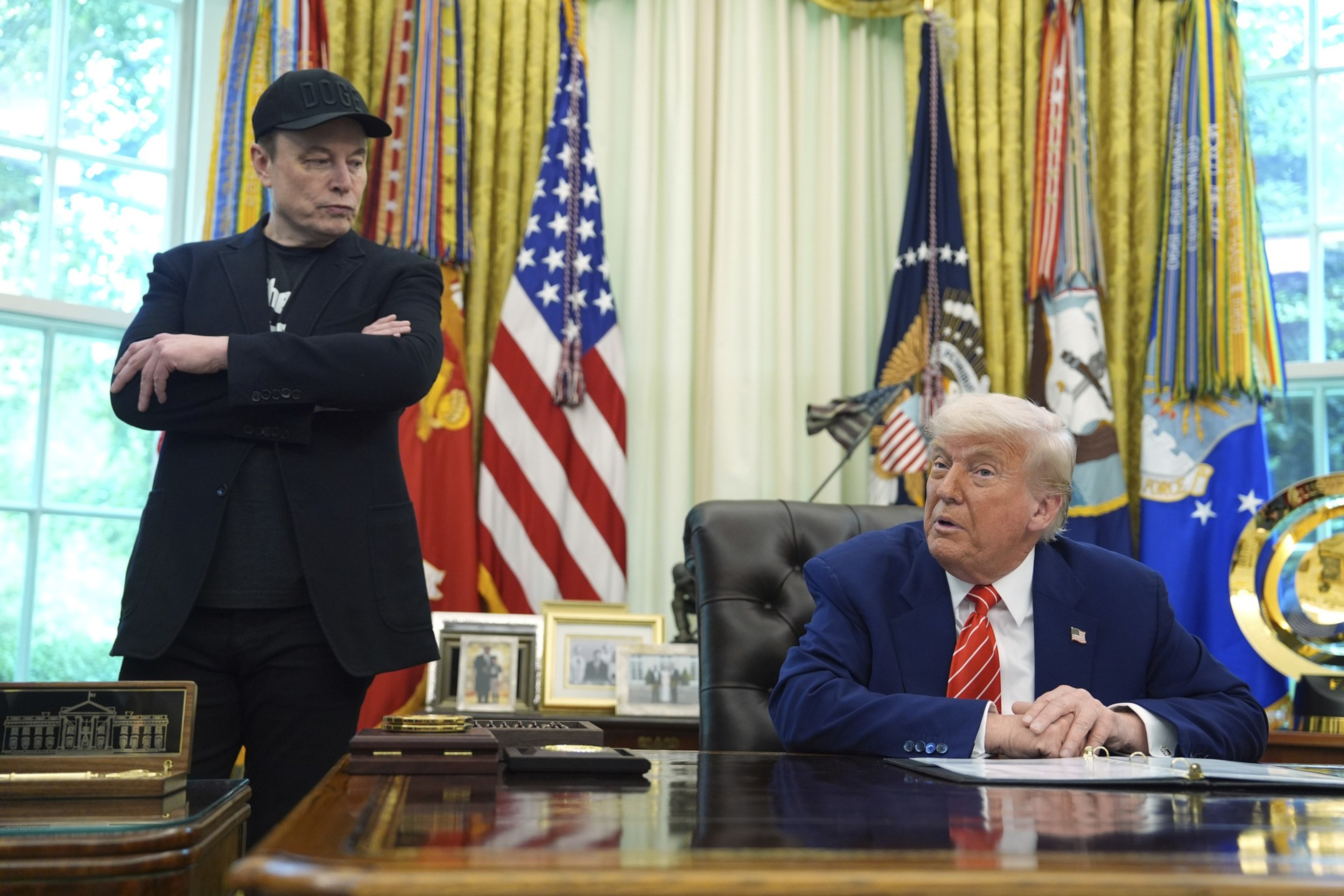 A man in a black cap and jacket stands with arms crossed near another man in a blue suit seated at a desk. Flags and ornate decorations are visible behind them.