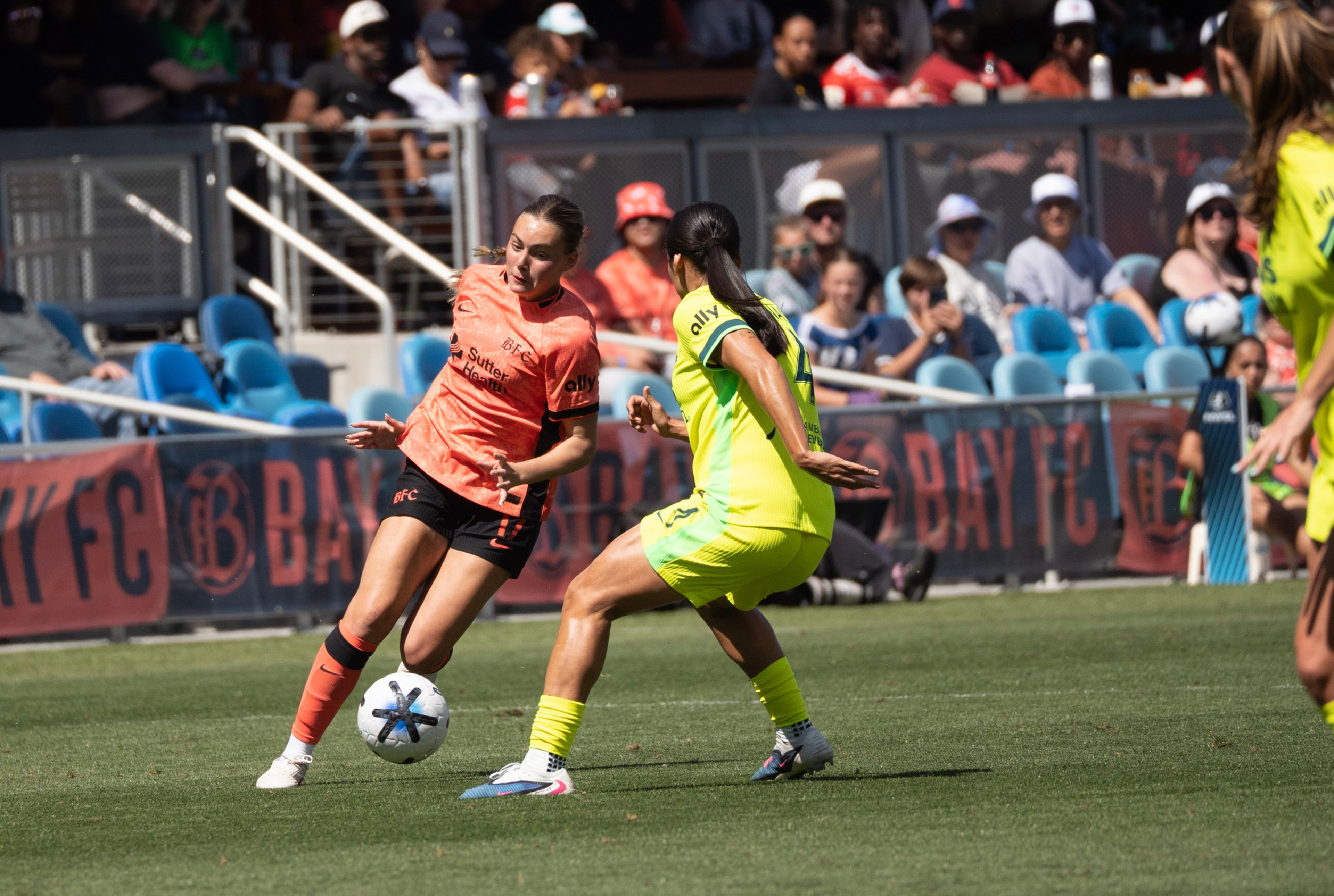 A soccer player in an orange jersey dribbles the ball while a player in a yellow jersey defends on a sunny field with spectators in the background.